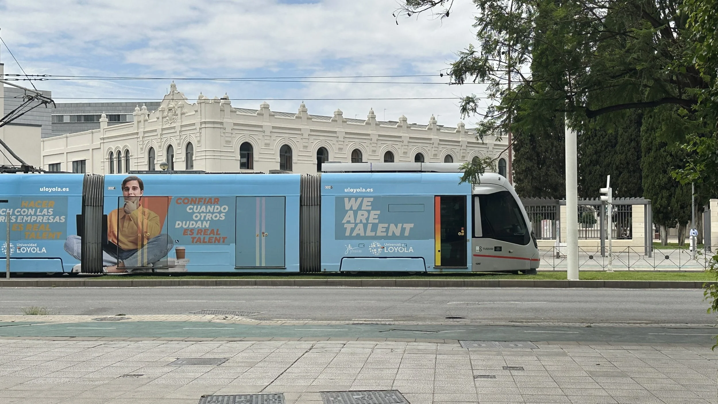Un tranvía azul con anuncios promocionales de la Universidad Loyola, en una calle con árboles y un edificio antiguo de fondo.