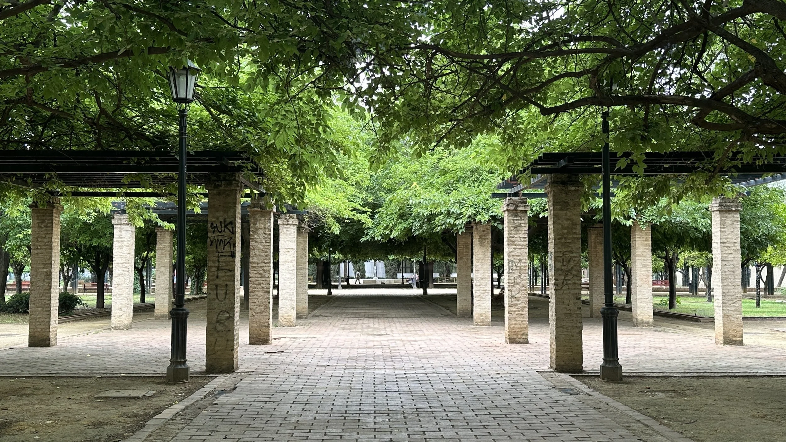 Un parque con pasillo pavimentado, columnas de ladrillo, árboles con hojas verdes frondosas y farolas en la sombra.