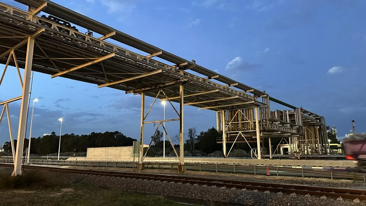 Infraestructura de tuberías y estructuras metálicas sobre una carretera, con luces y fondo de cielo azul y nubes.