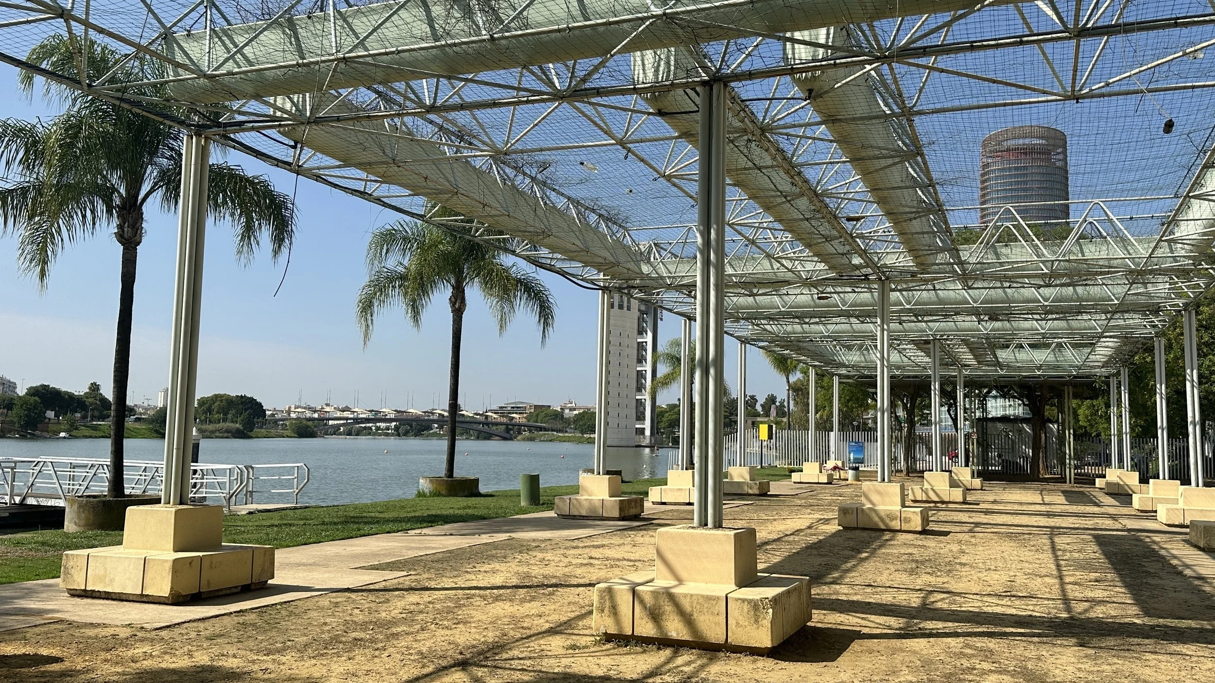 Estructura moderna de techumbre en una zona al aire libre junto a un río, con palmeras, bancos de concreto, árboles y una ciudad en el fondo.