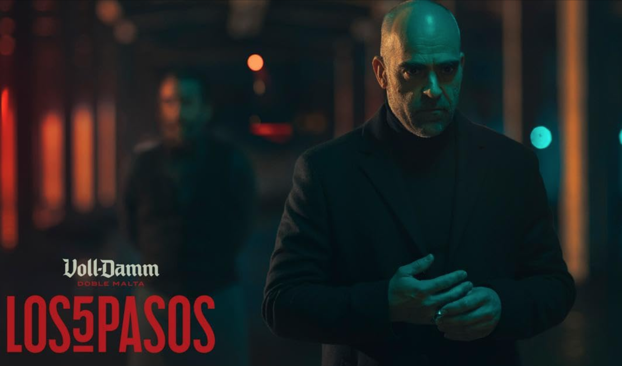 Hombre calvo con traje negro en una calle nocturna iluminada con luces de neón, sosteniendo un cigarro, con texto en la esquina inferior izquierda que dice 'Volk-Damm Doble Malta Los Pasos'