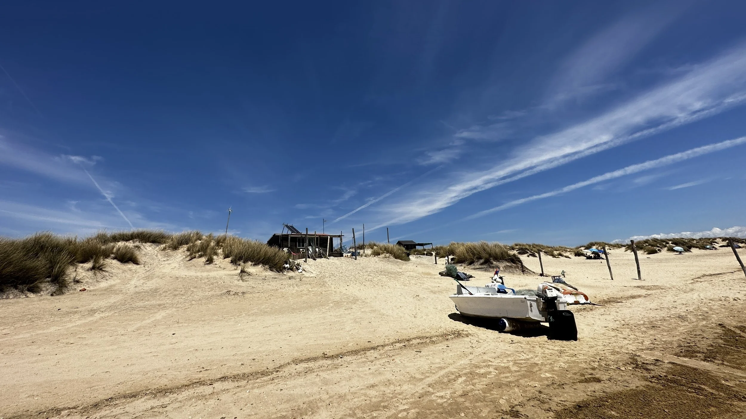Playa arenosa con vegetación de dunas, casas de playa y botes en la arena bajo un cielo azul con nubes delgadas.