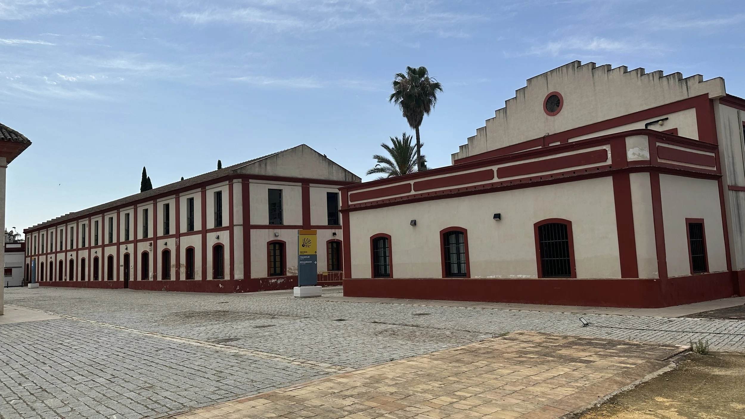 Edificio blanco con detalles rojos y ventanas con barras, en una plaza con pavimento de piedra, hay palmeras y un cielo despejado.