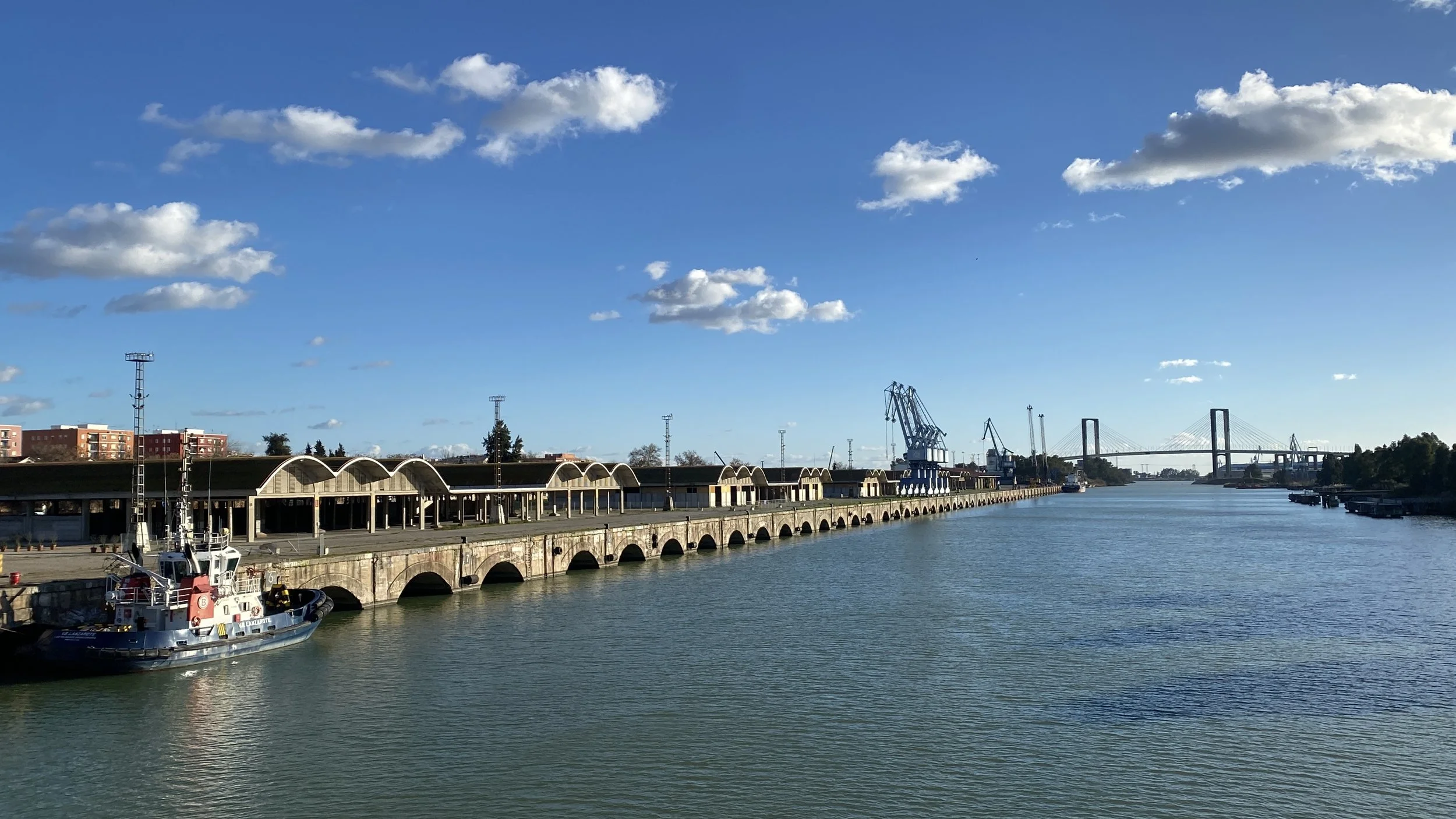 Vista de un puerto con barcos y un puente en la distancia, cielo despejado con algunas nubes.