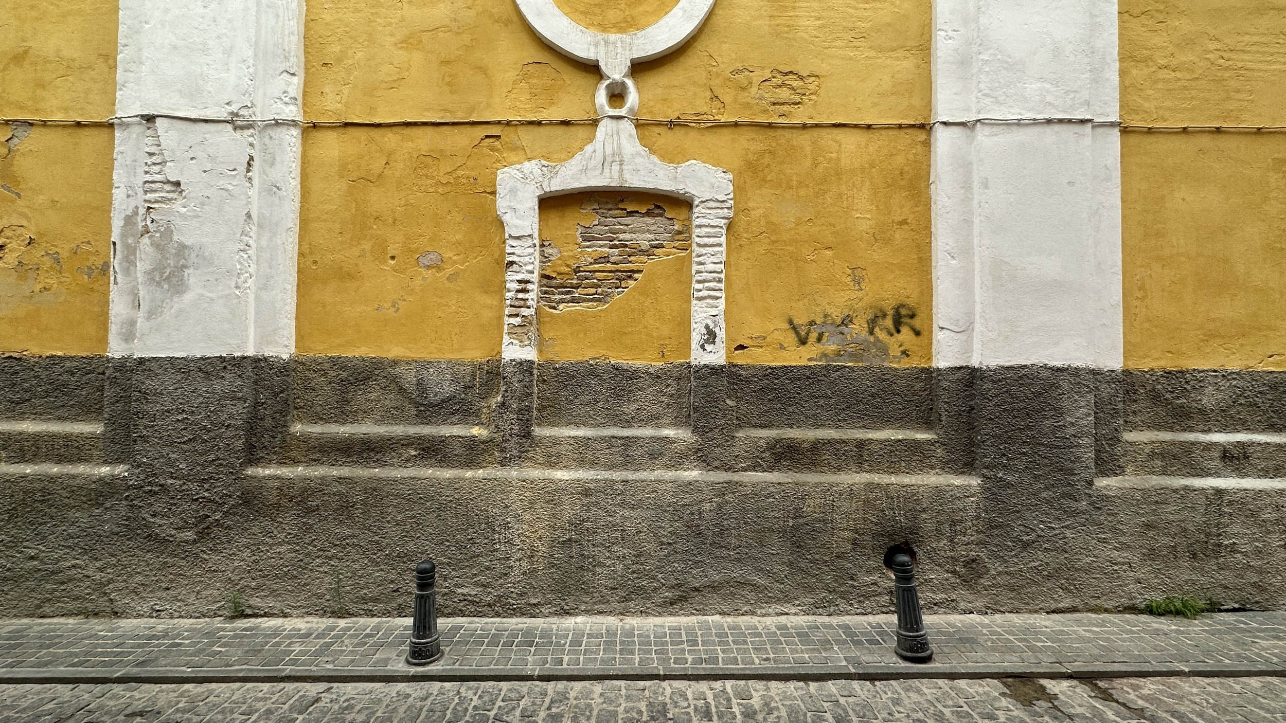 Una pared amarilla con detalles blancos y una sección de ladrillos expuestos, que parece estar dañada. En la parte inferior, una base de concreto gris y un suelo de piedra. Dos bolardos negros de metal en primer plano.