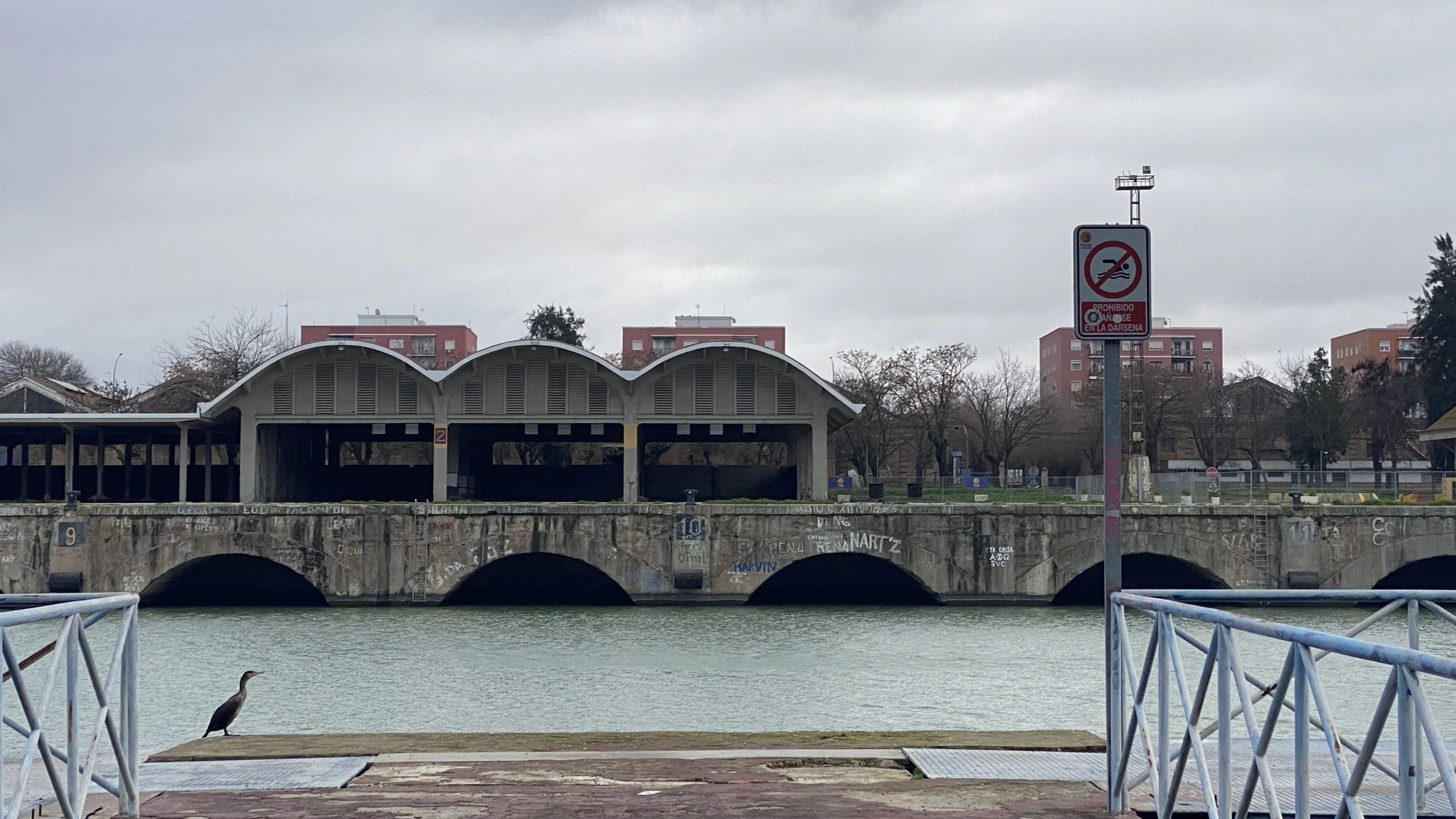 Puente de piedra sobre el río con estructuras de techo curvas y un cartel de prohibido bañarse en la Dársena