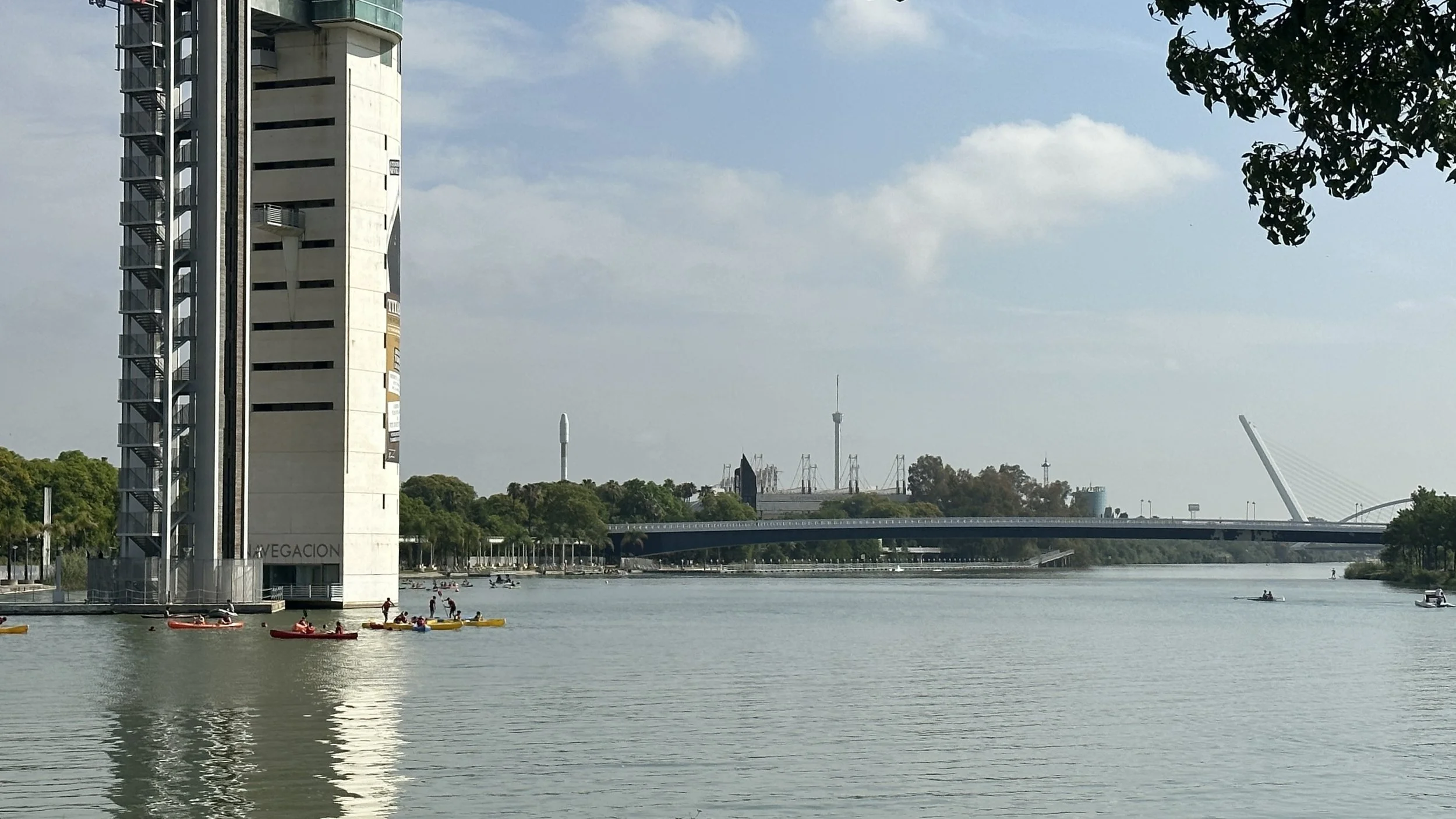 Edificio junto al río con personas remando en kayak y kayak, puente y ciudad en el fondo.