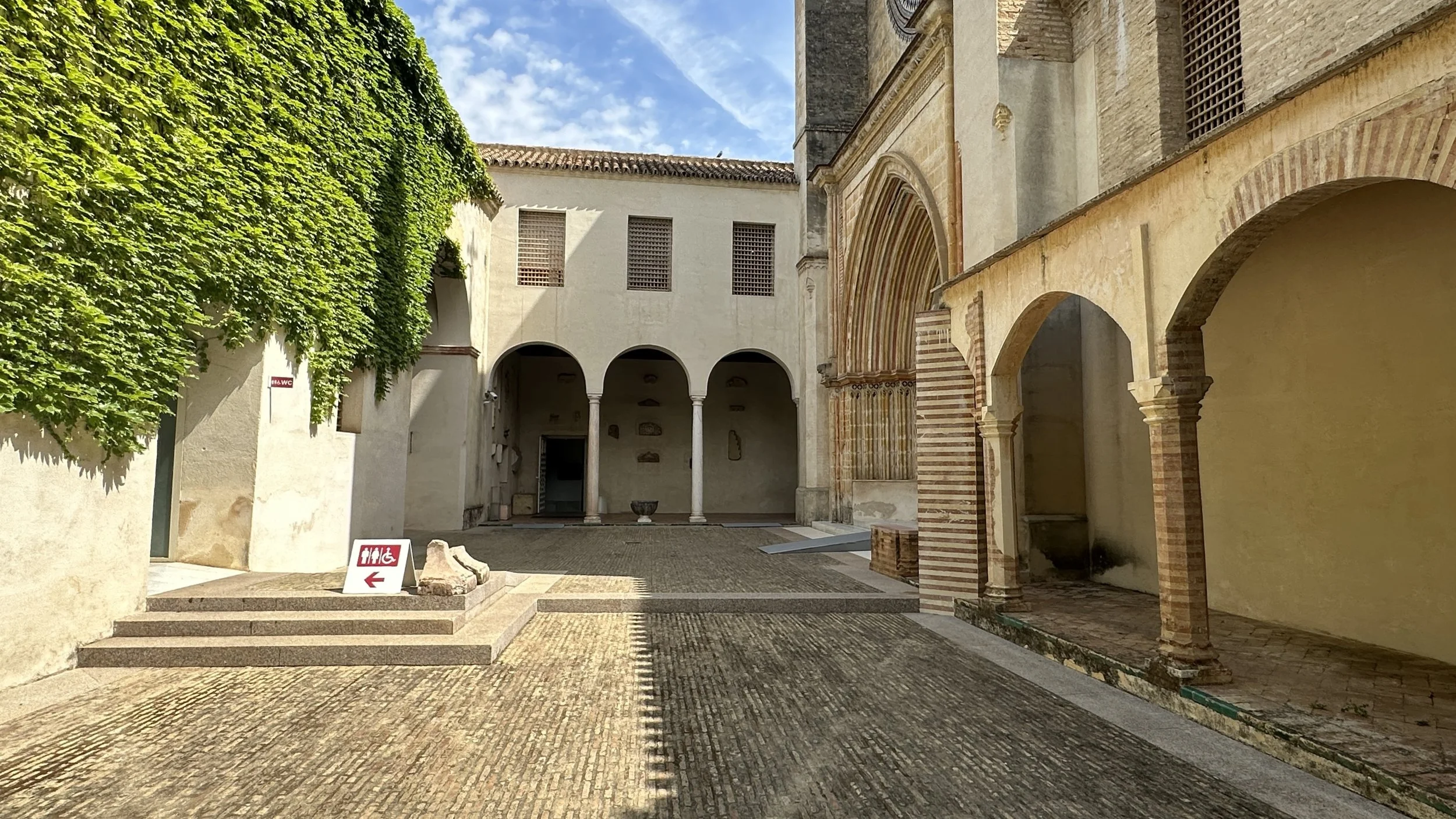 Patio de un edificio histórico con arcos, paredes de piedra, vegetación en un lado y señalización de servicios en el suelo.