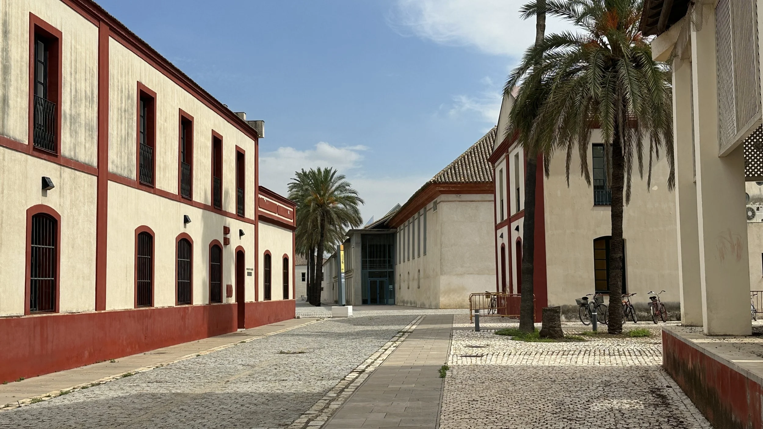 Una plaza con edificios de colores beige y rojo, palmeras y bicicletas estacionadas, cielo despejado.