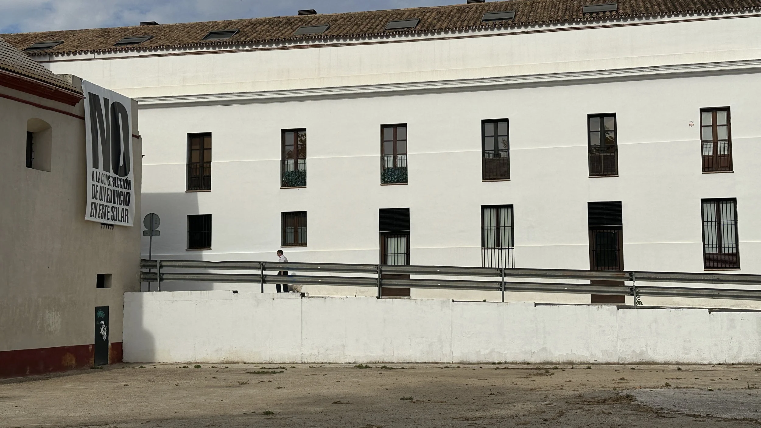 Edificio de color blanco con ventanas y puertas de metal, en la calle con una persona caminando y un perro pequeño. Cartel grande en la parte izquierda que dice ‘NO a la construcción de un edificio en este solar’.
