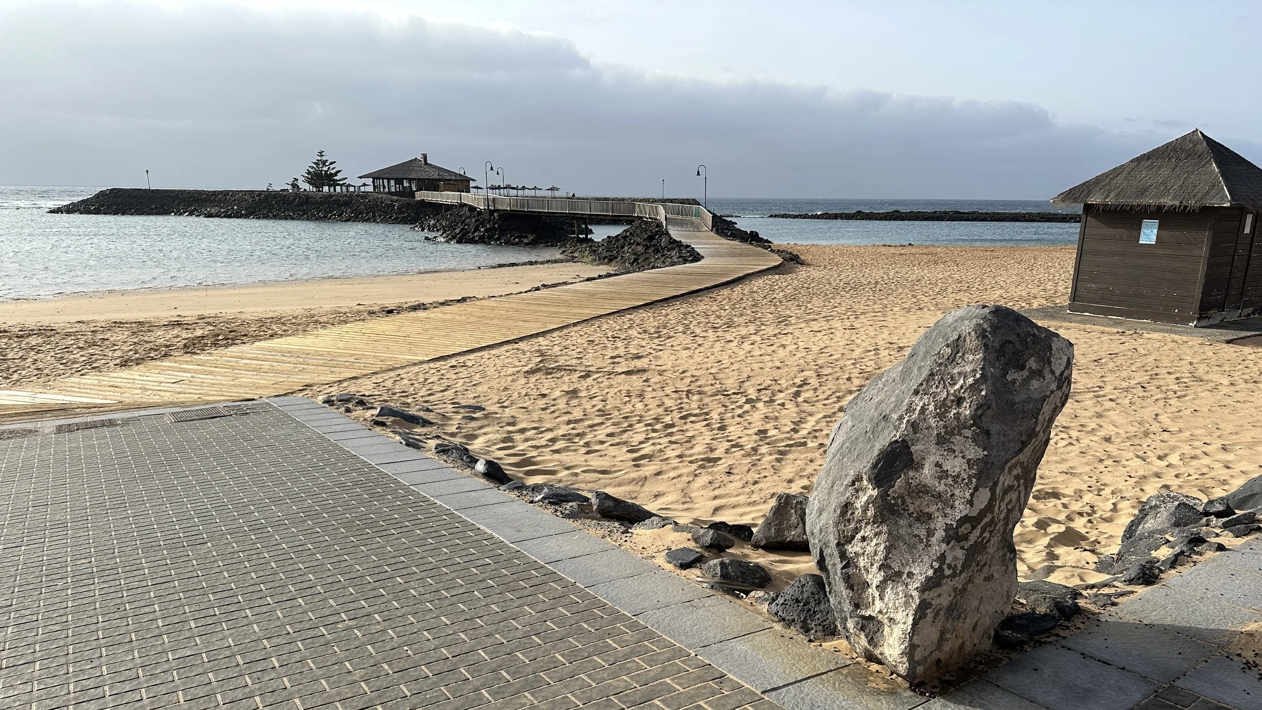 Camino de madera que atraviesa la playa, rocas grandes en primer plano, cabañas y un muelle en el fondo, con cielo nublado.