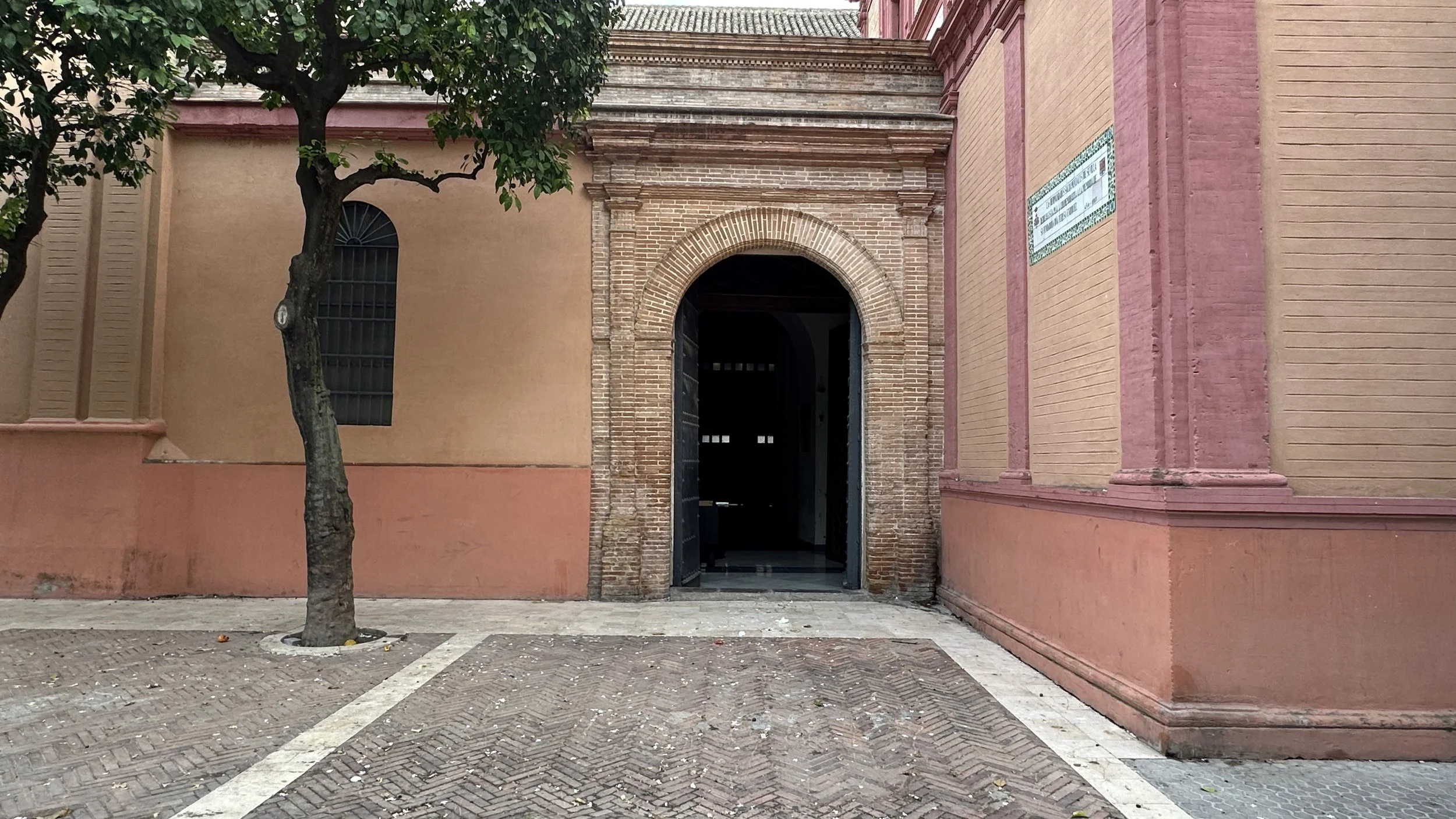 fachada de una iglesia con arco en su entrada, paredes de color rosa y ladrillo, y un árbol en la acera delante de la entrada.