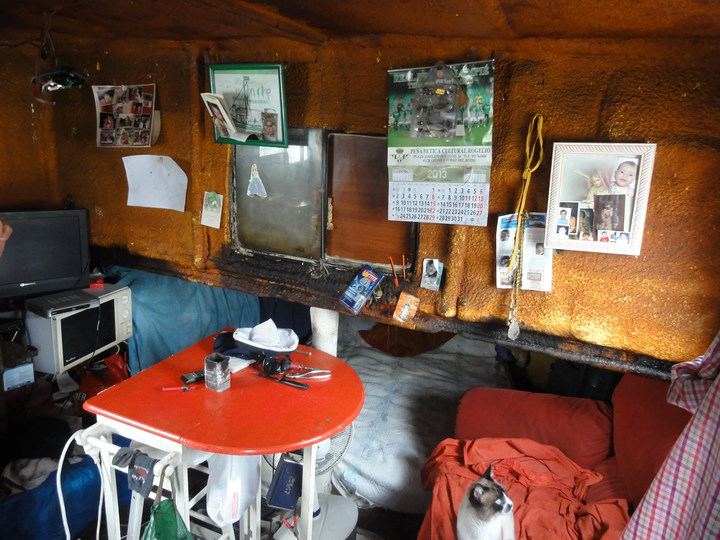 Interiores de una casa con paredes rústicas de color marrón. En la parte izquierda hay una televisión pequeña sobre una mesa con algunos objetos. En la mesa tiene herramientas y objetos relacionados con trabajos manuales como brocas, un cubo de alumi