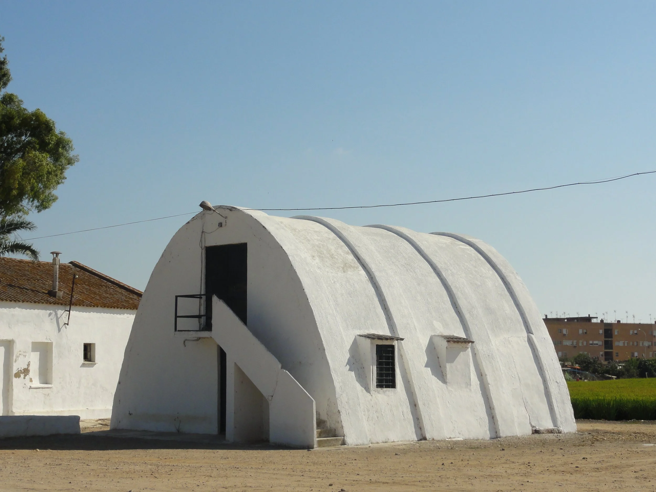 Construcción blanca con forma de arcos, ubicada en un área abierta, con cielo despejado y algunos árboles en el fondo.