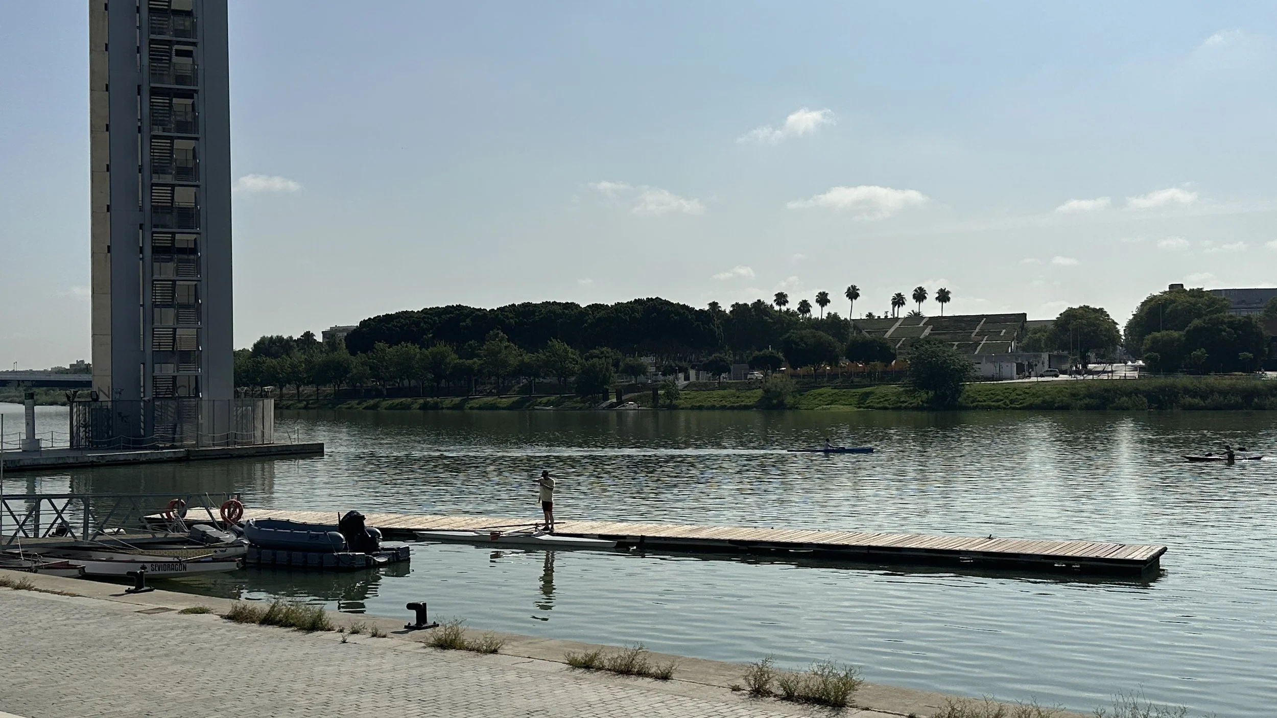 Un paso peatonal, un muelle con botes, gente en el río, árboles y una estructura moderna junto al agua, con palmeras y vegetación en el fondo.