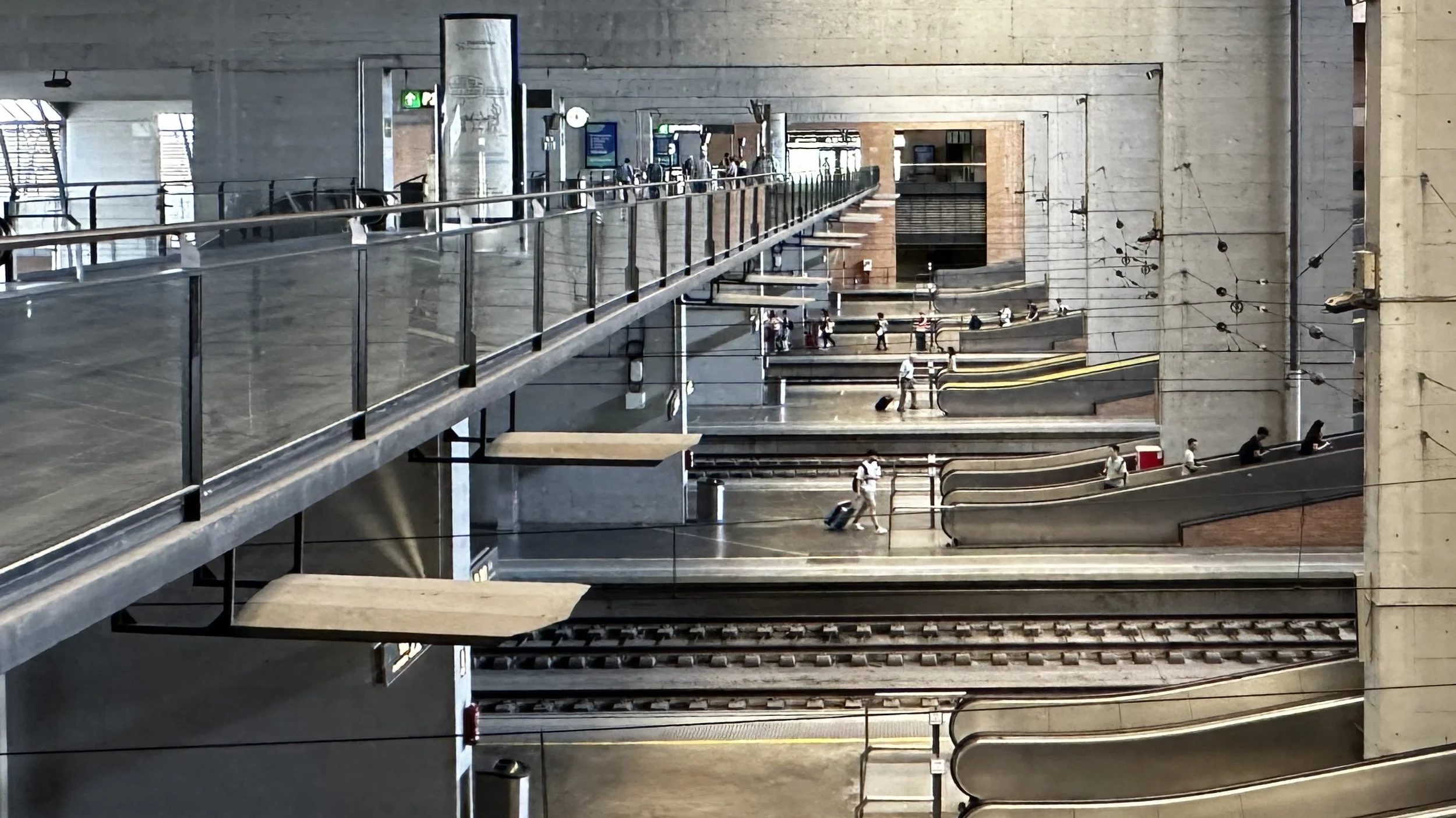 Interior de una estación de tren con varias plataformas, escaleras y pasajeros con equipaje.