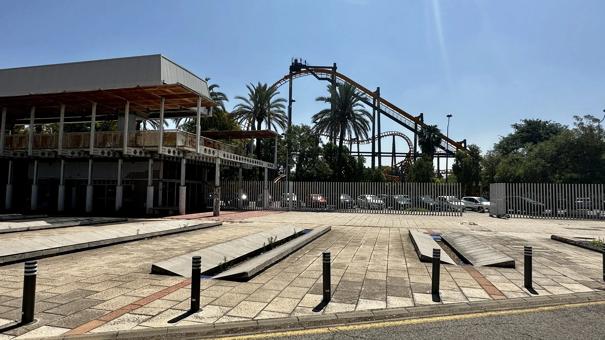 Una calle con un parque de diversiones al fondo, con montañas rusas y árboles de palma, y estacionamiento con autos, en un día soleado.