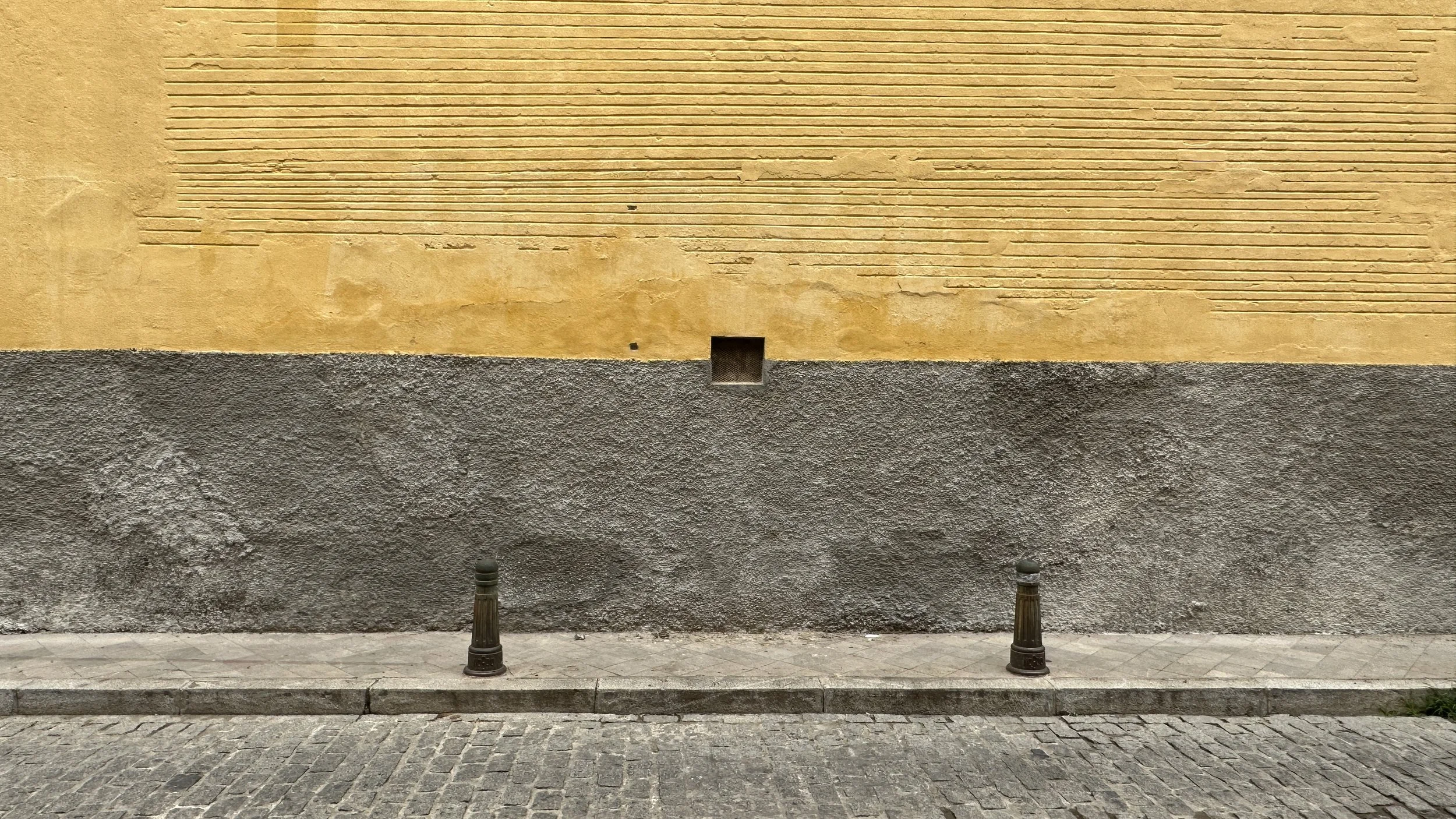 Muro de pared con textura diferente en la parte superior y inferior, en la parte central hay una pequeña abertura cuadrada y en la acera hay dos bolardos