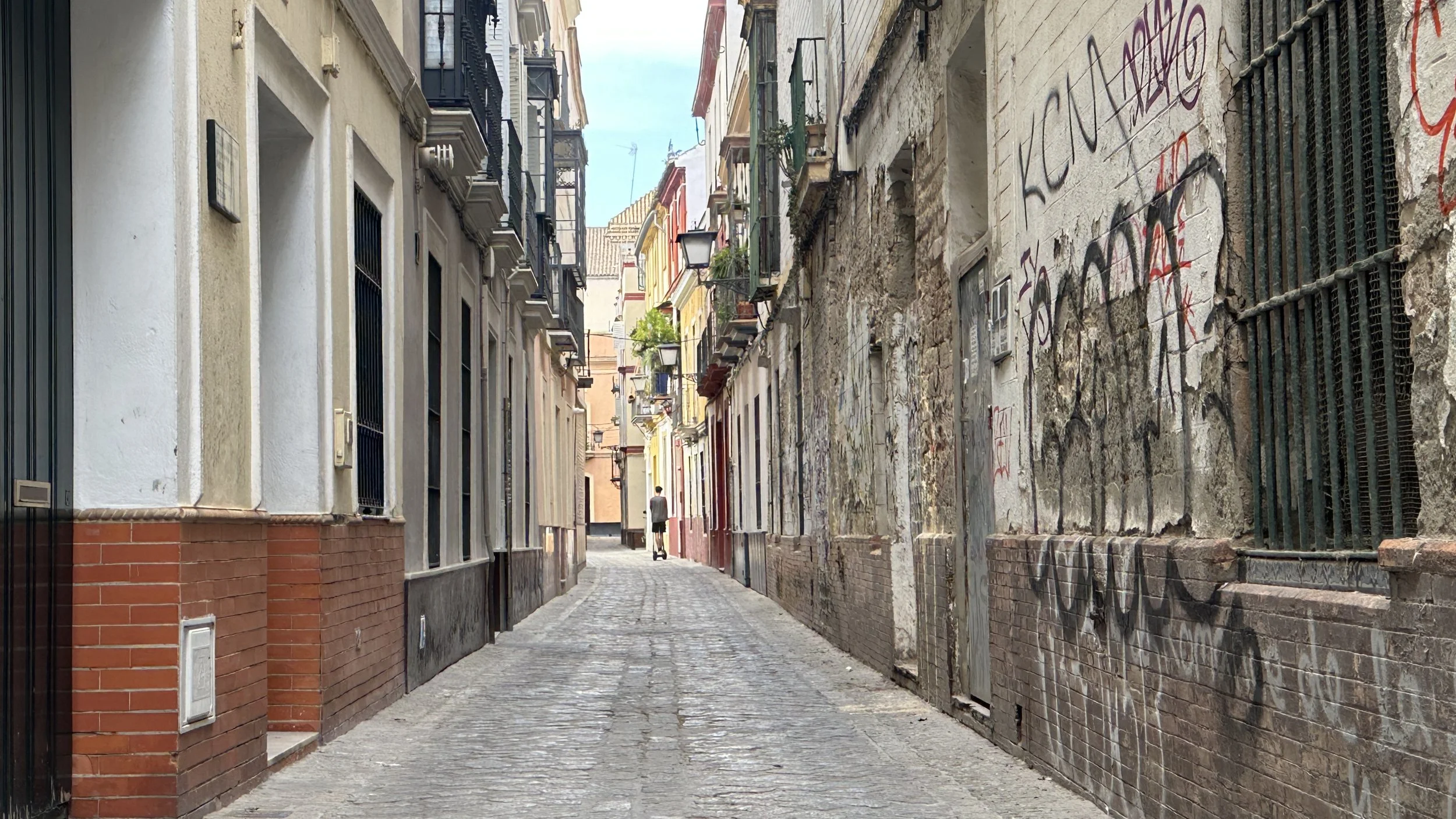 Calles estrechas entre edificios en una ciudad, con paredes envejecidas y graffitis. Un peatón camina en la distancia, cielo despejado visible.