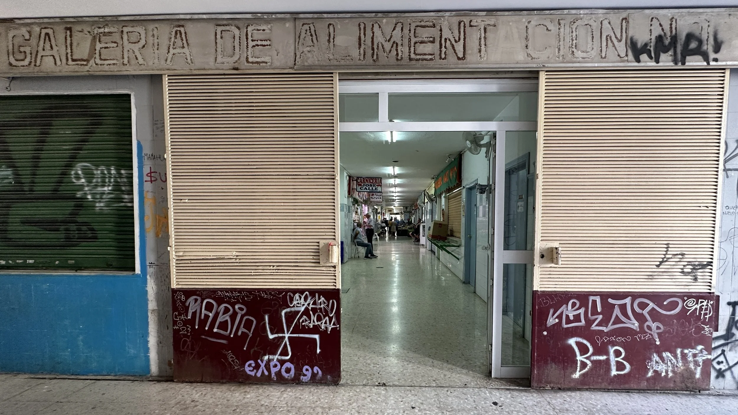 Vista del interior de un centro comercial con pasillo y varias tiendas, algunos clientes sentados y otros caminando, con cartelera y graffitis en las paredes.