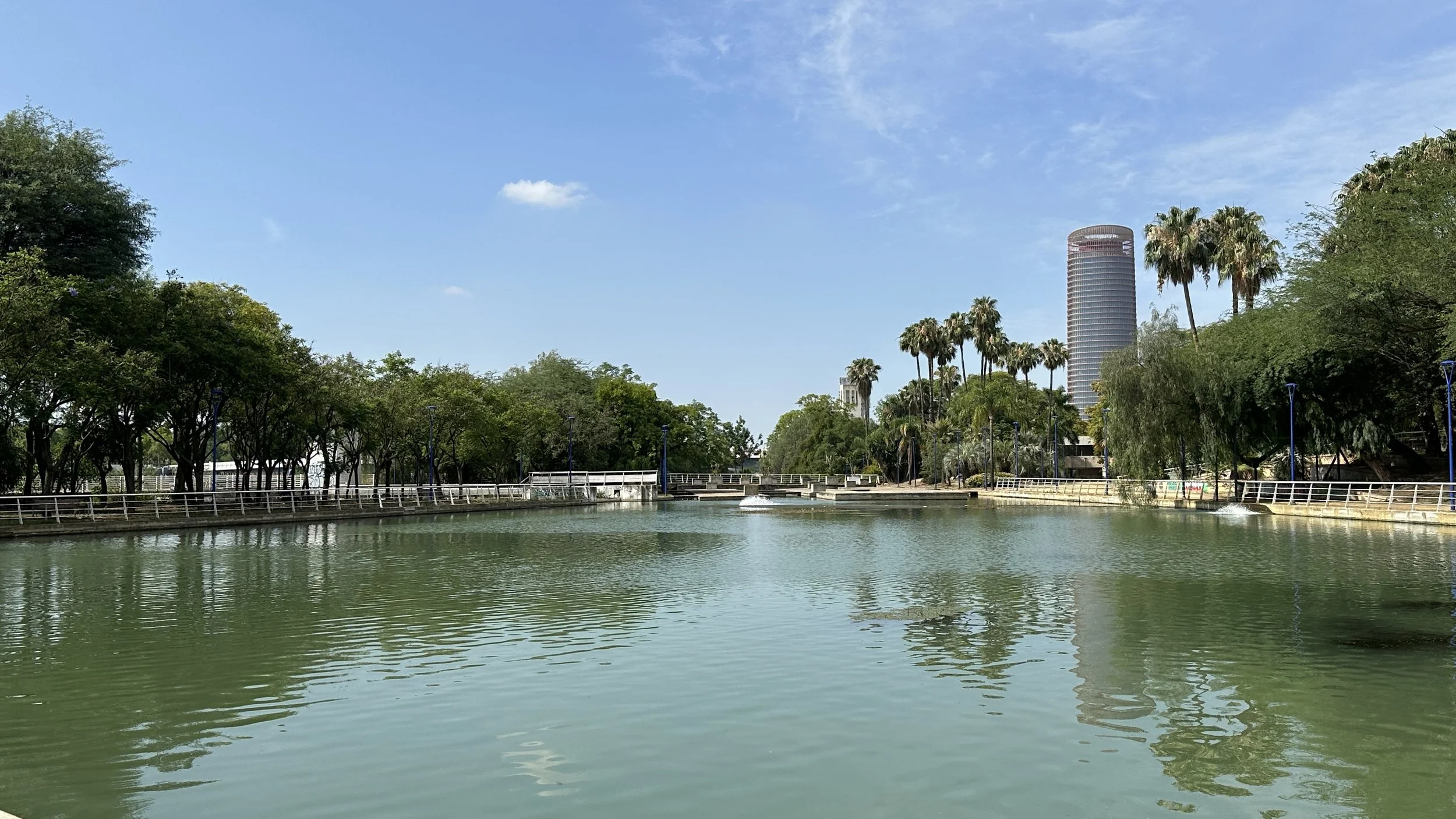 Un parque con árboles y un cuerpo de agua, con una ciudad en el fondo que incluye un edificio alto cilíndrico.