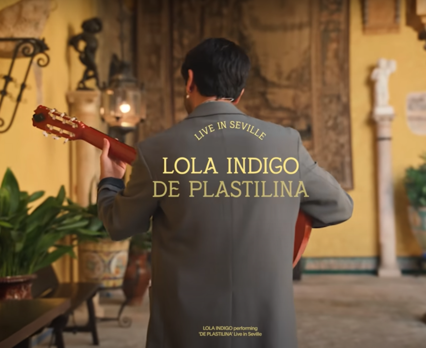 Una mujer vista de espaldas, tocando la guitarra en un entorno decorado con objetos antiguos y plantas, con texto en la imagen que dice: 'Vive en Sevilla. Lola Indigo de Plastilina'.