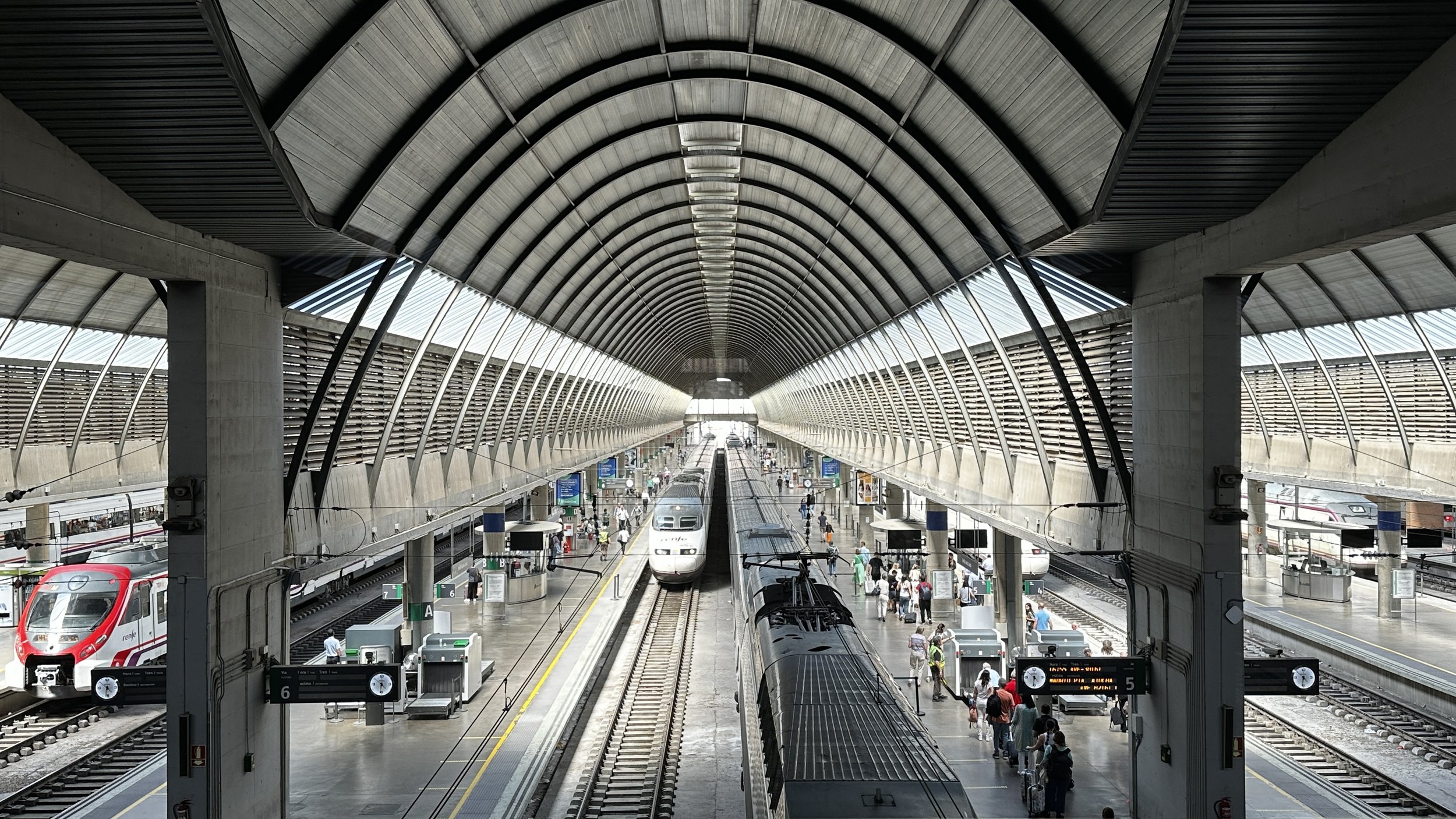 Estación de tren moderna con techos arqueados de metal, trenes en las vías y pasajeros esperando en la plataforma.