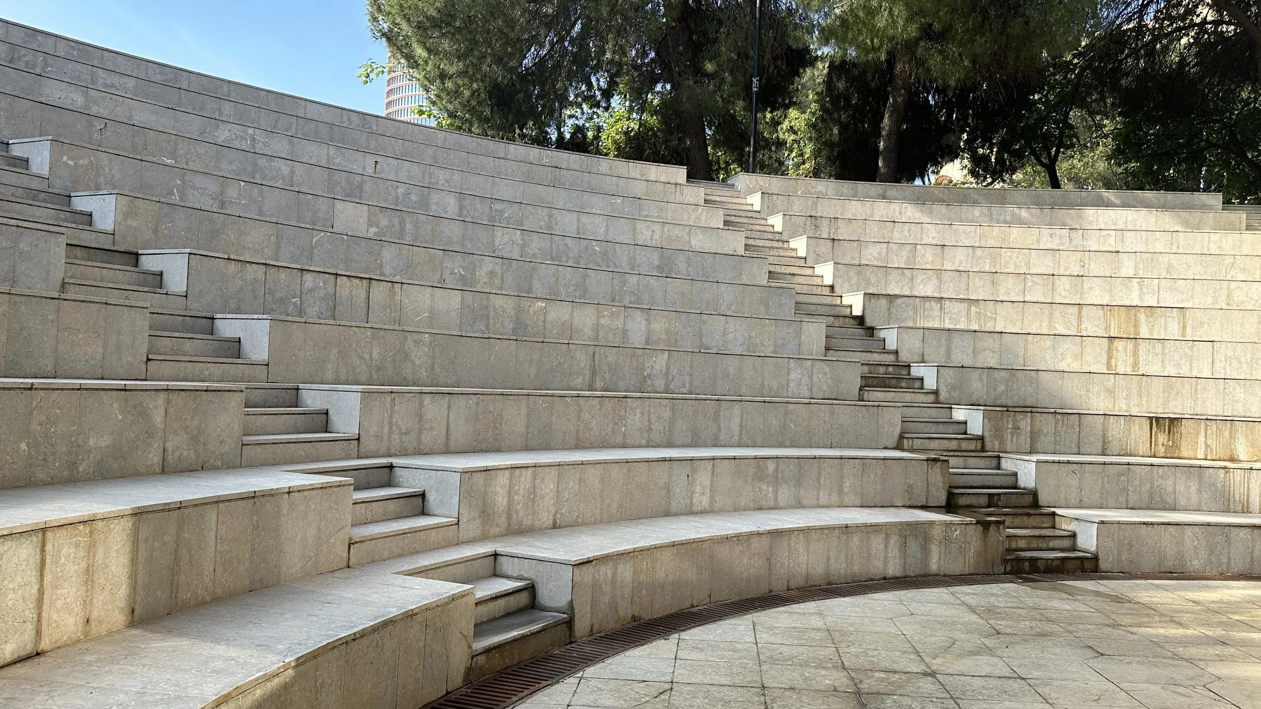 Escalinatas de piedra en un espacio público al aire libre, con árboles en el fondo y cielo azul.