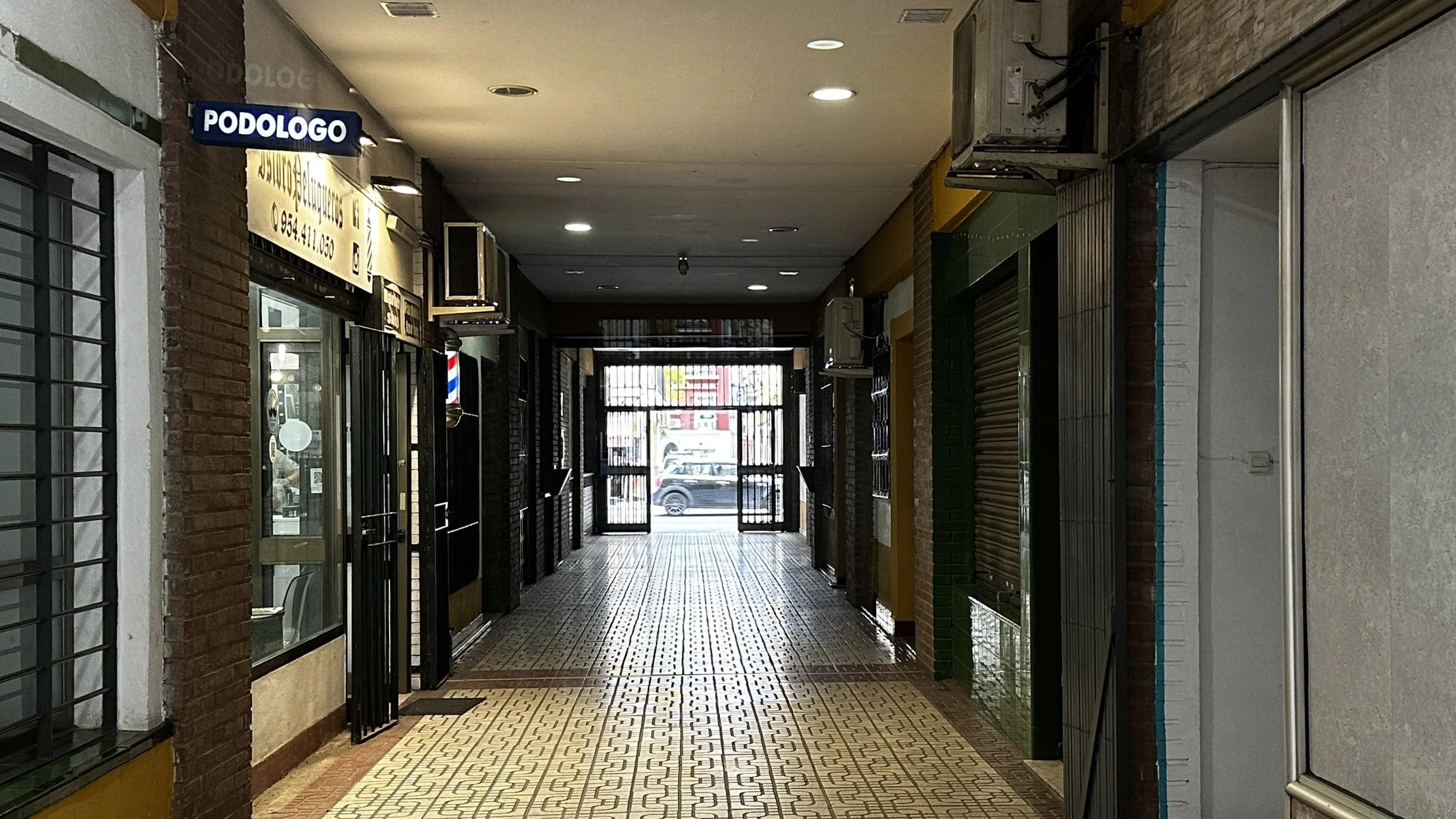 Pasillo interior con tiendas cerradas, puertas metálicas con rejas, piso con baldosas decorativas y señal de 'Podólogo' en letrero en la parte superior izquierda.