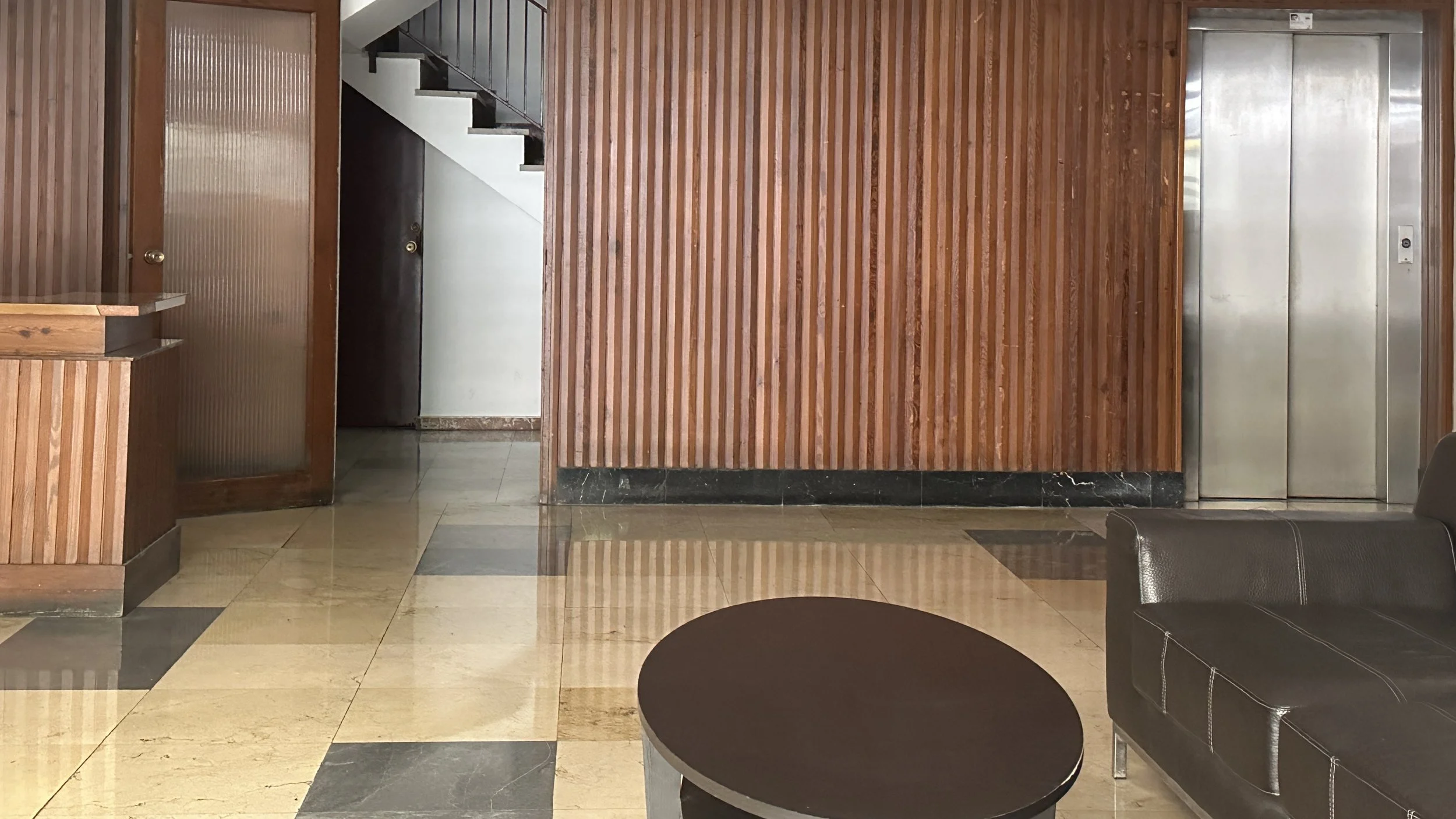 Interior de un edificio con pared de madera, ascensor y área de descanso con sillas y mesas.