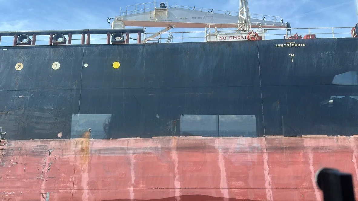 Fotografía de un buque en el puerto, con la parte inferior de la embarcación visible y un cielo despejado.