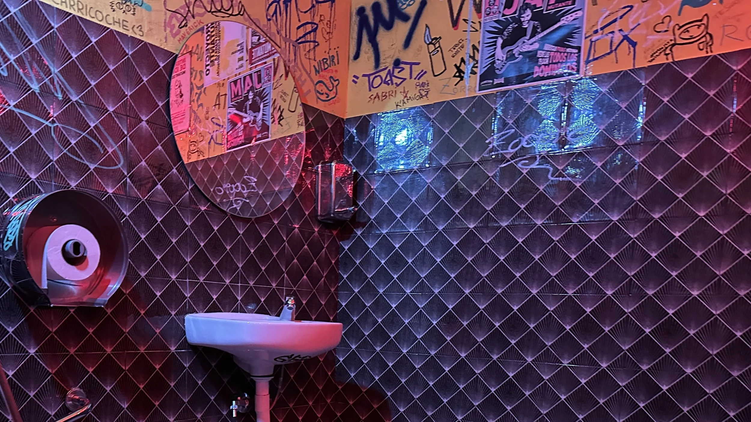 Cuarto con paredes decoradas con graffiti y posters musicales, espejo, lavabo, dispensador de jabón, y sistema de iluminación con luces de colores.