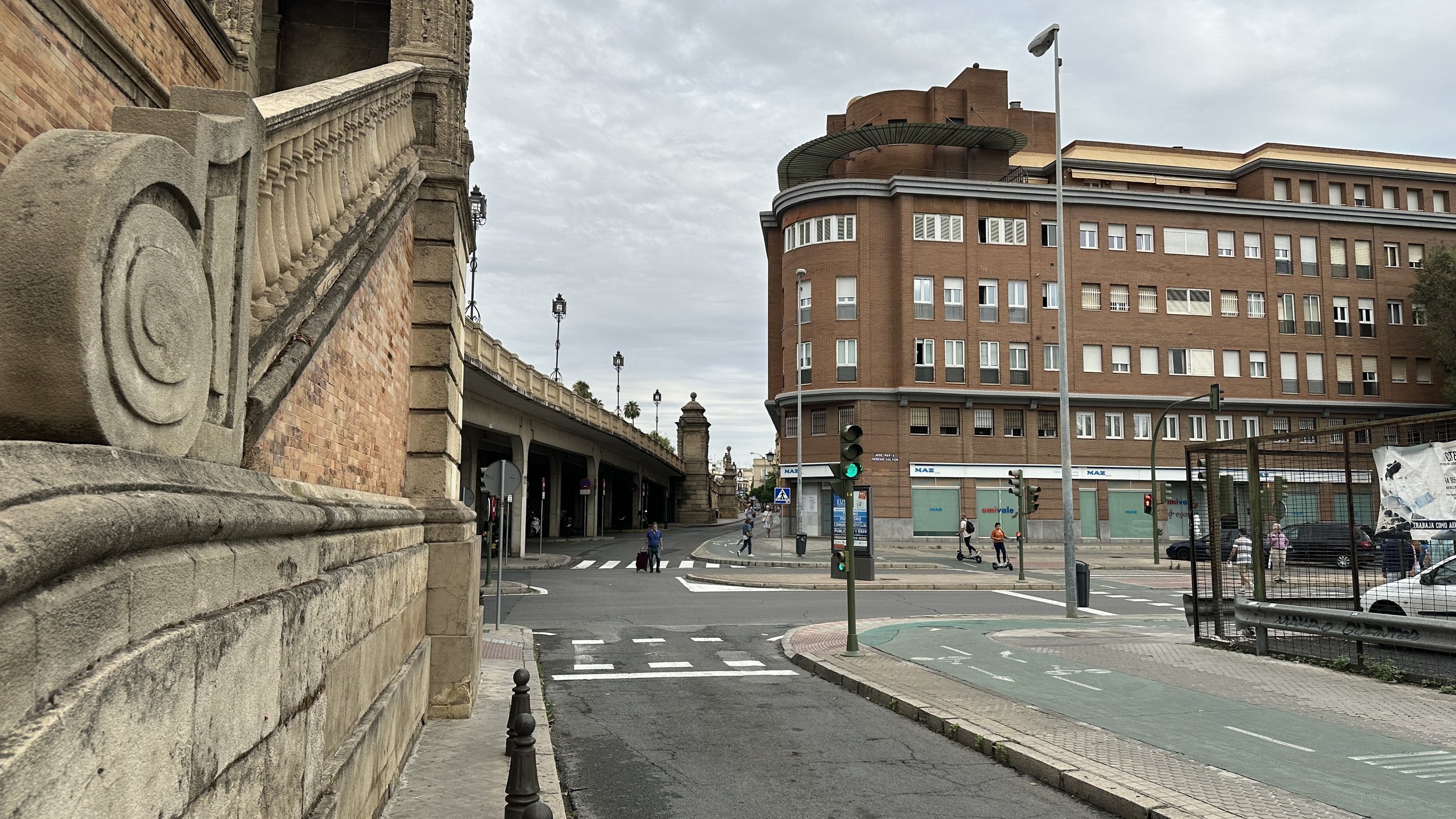 Vista urbana con un puente, un edificio grande de ladrillos, semáforos, personas cruzando y un cielo nublado.