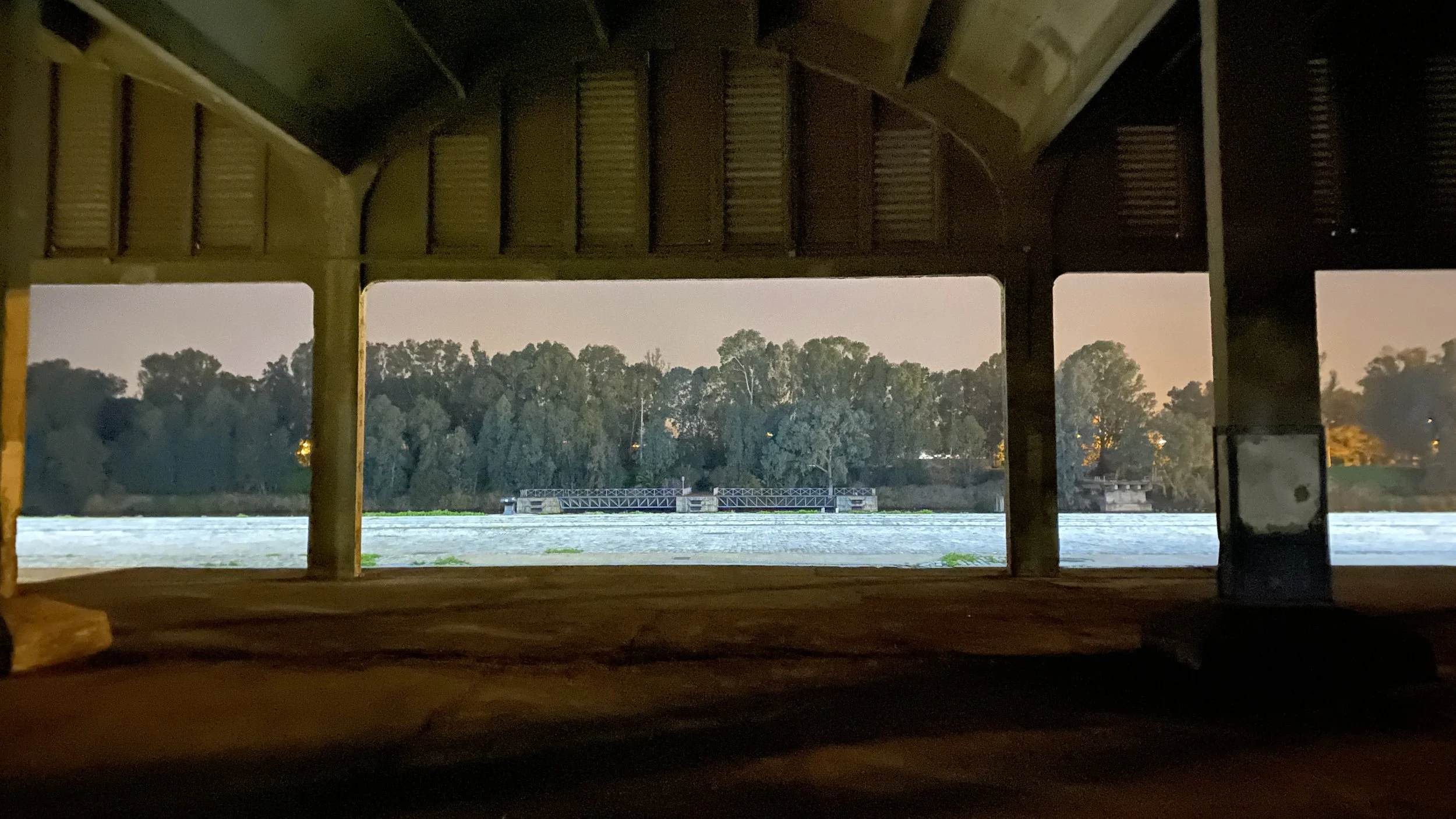 Vista de un río desde una estructura de madera, con árboles y un puente en el fondo, durante el anochecer.