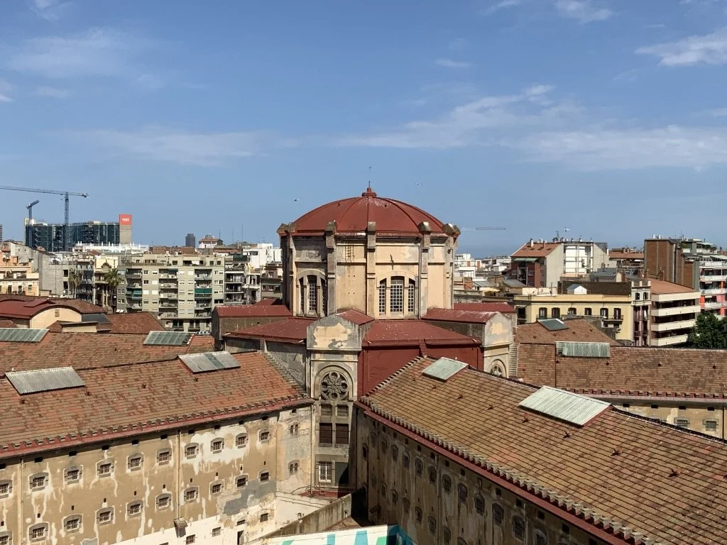 Vista panorámica de una ciudad con edificios modernos y antiguos, en el centro una iglesia con cúpula roja y fachada de piedra, bajo un cielo parcialmente nublado.
