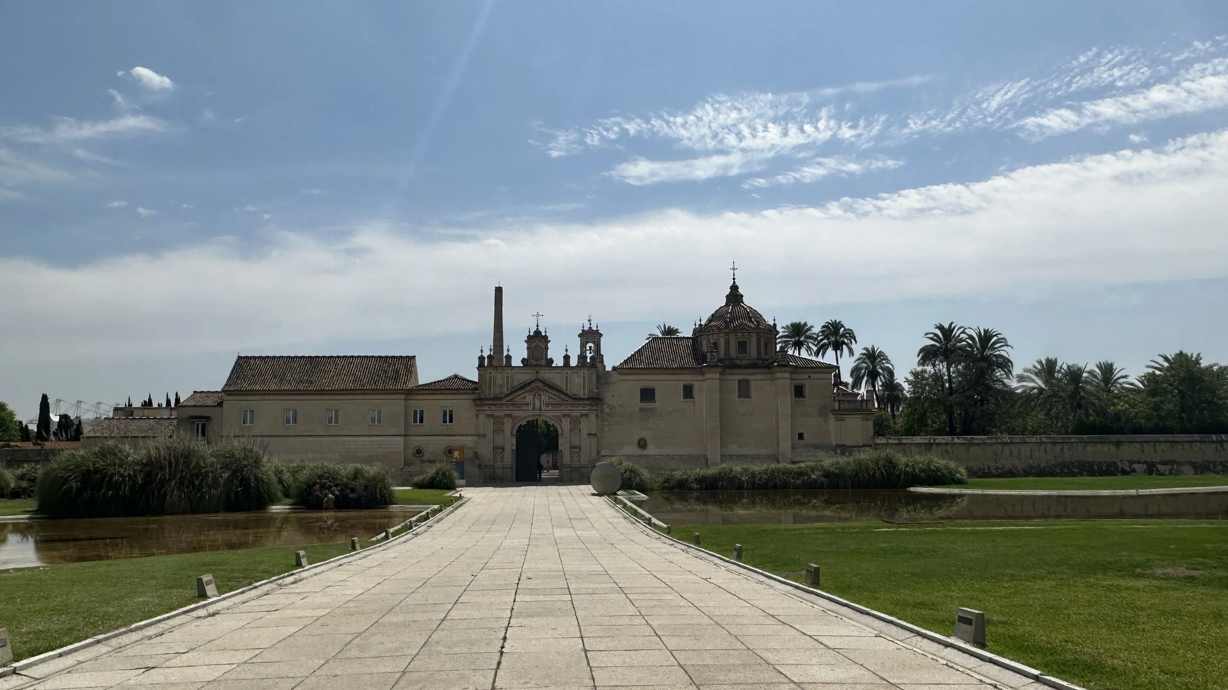 Una vista de un edificio histórico con paredes beige, una puerta de entrada arqueada, varias torres y un campanario, rodeado de árboles de palma y un jardín con camino de piedras.