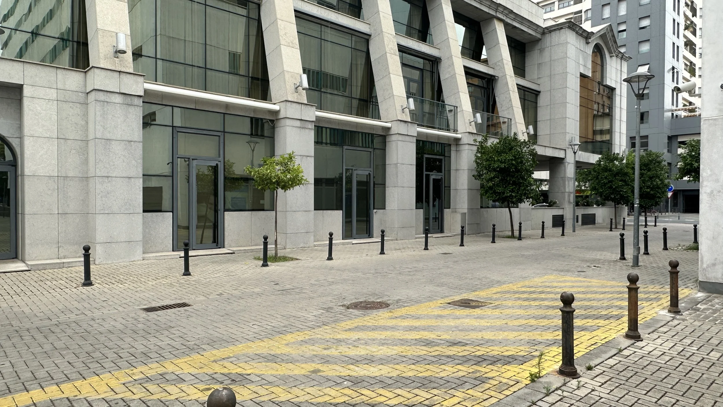 calle en una ciudad con edificios modernos de vidrio y concreto, árboles pequeños, postes y un piso de adoquines