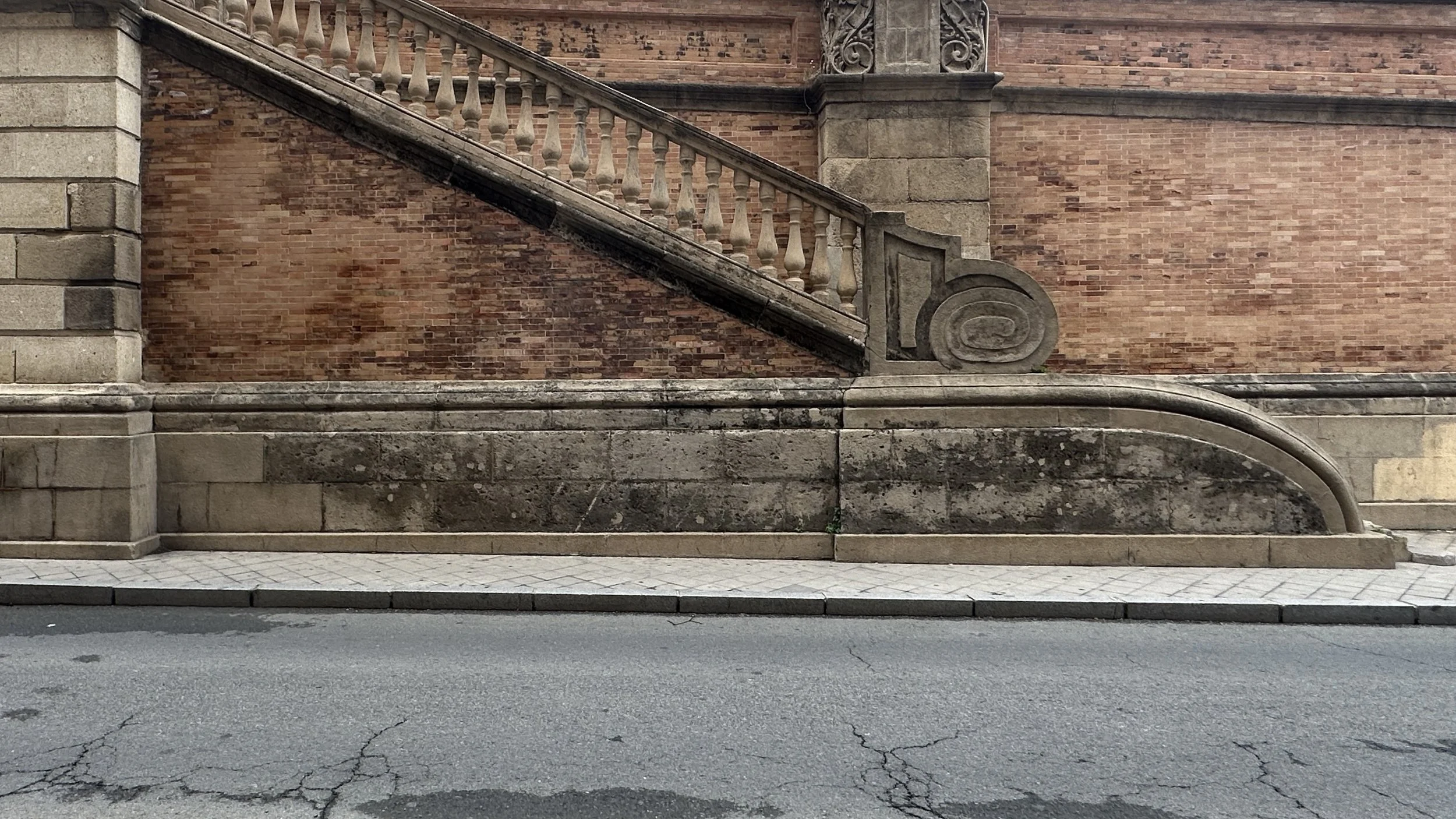 Escalera de piedra con barandilla de balustrada y pared de ladrillos en el fondo, frente a la calle pavimentada.