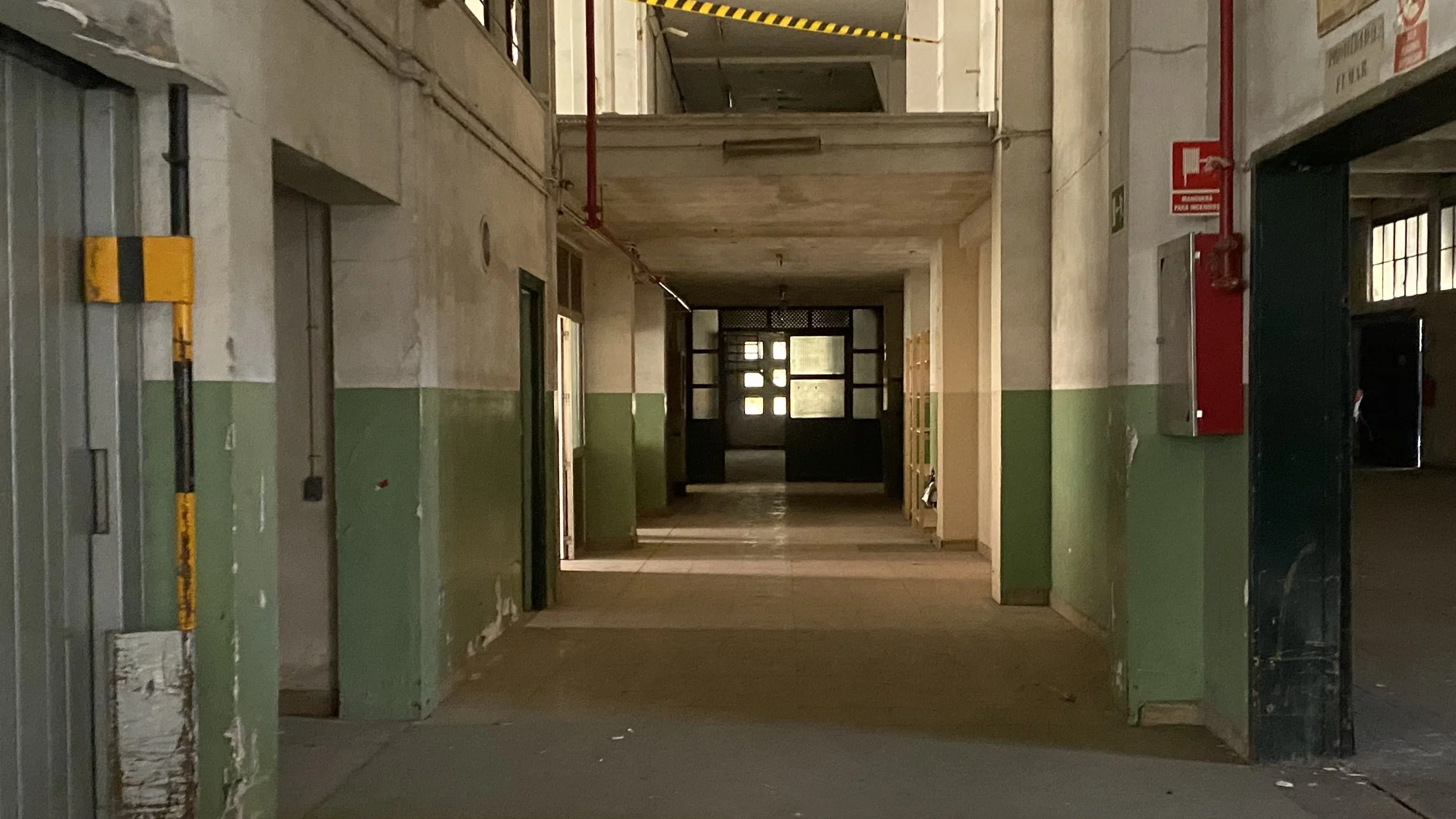 Pasillo vacío en un edificio escolar o industrial con paredes parcialmente pintadas en verde y beige, puertas abiertas, ventanas y un ventilador en el techo.