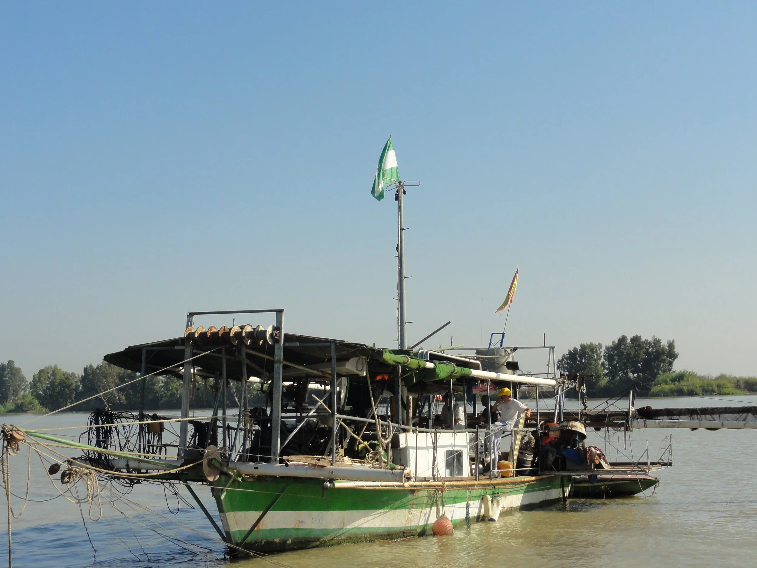 Barco de madera amarrado en río con bandera verde y blanca en mástil y personas en el muelle.