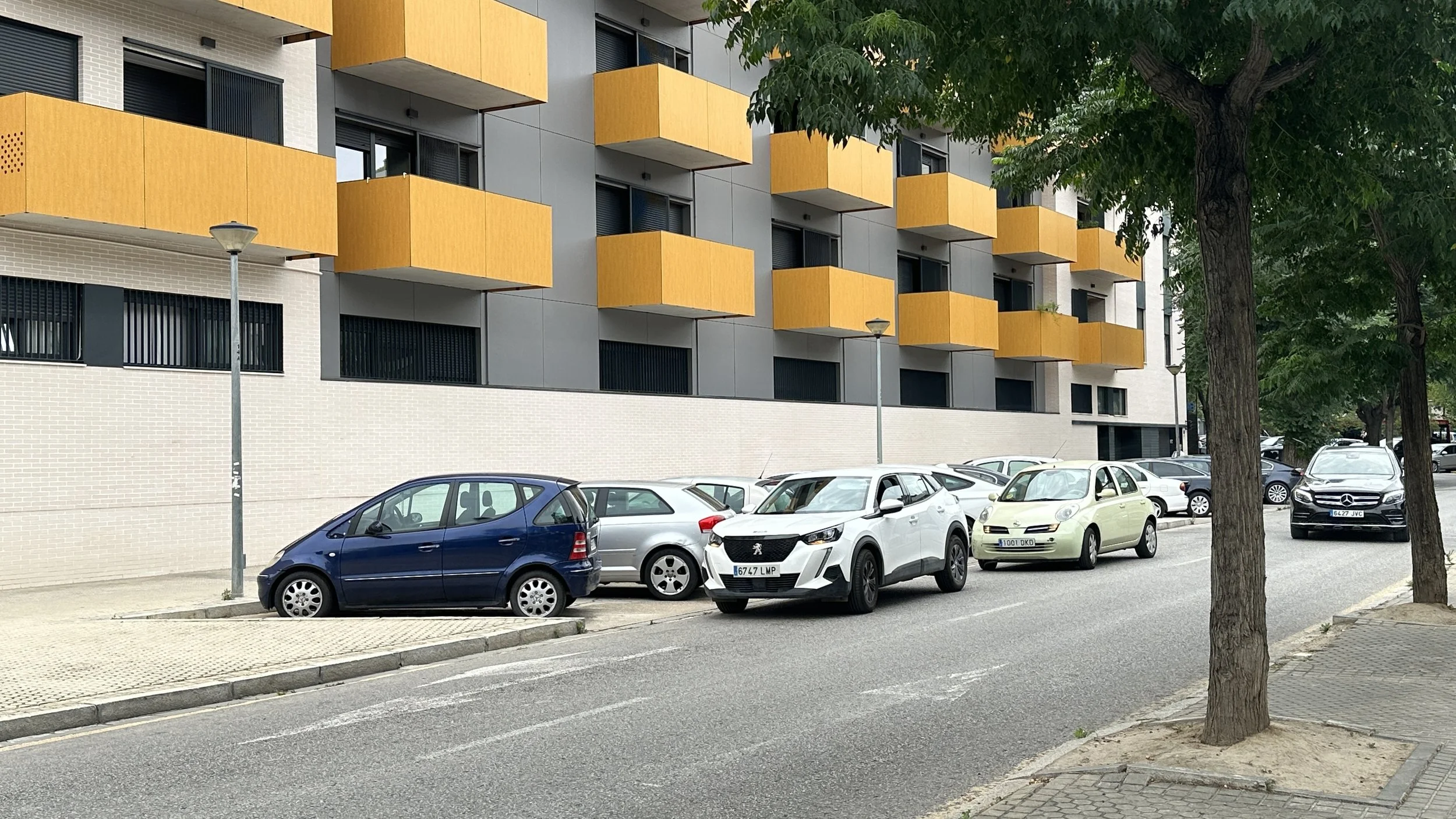 Estacionamiento con varios autos frente a un edificio moderno con balcones naranjas y ventanas negras, árboles a lo largo de la acera.