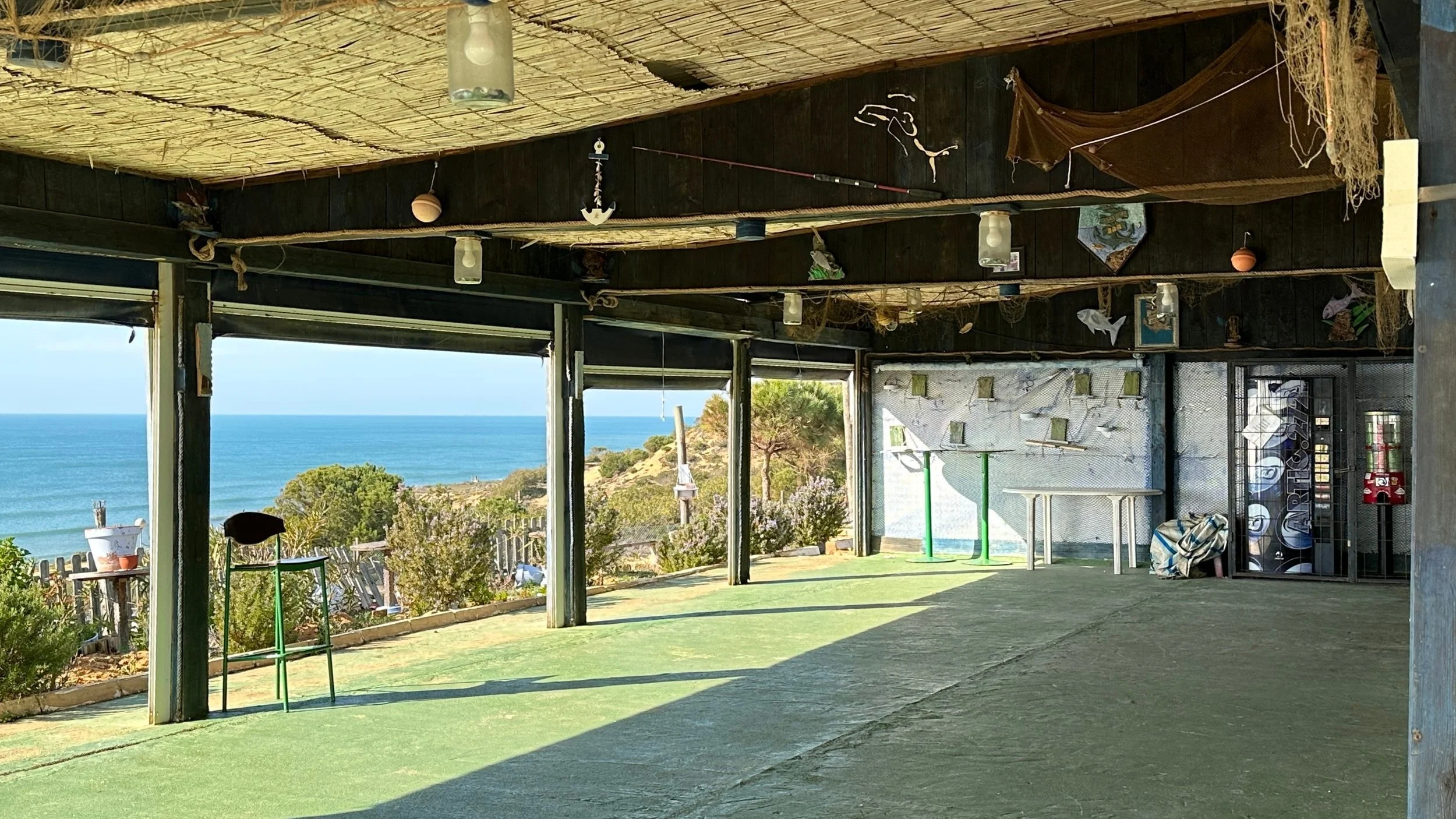 Vista de un espacio con techo de cañas y paredes de madera, con vista panorámica del mar y la playa en el fondo. Hay objetos decorativos y equipo de gimnasio.