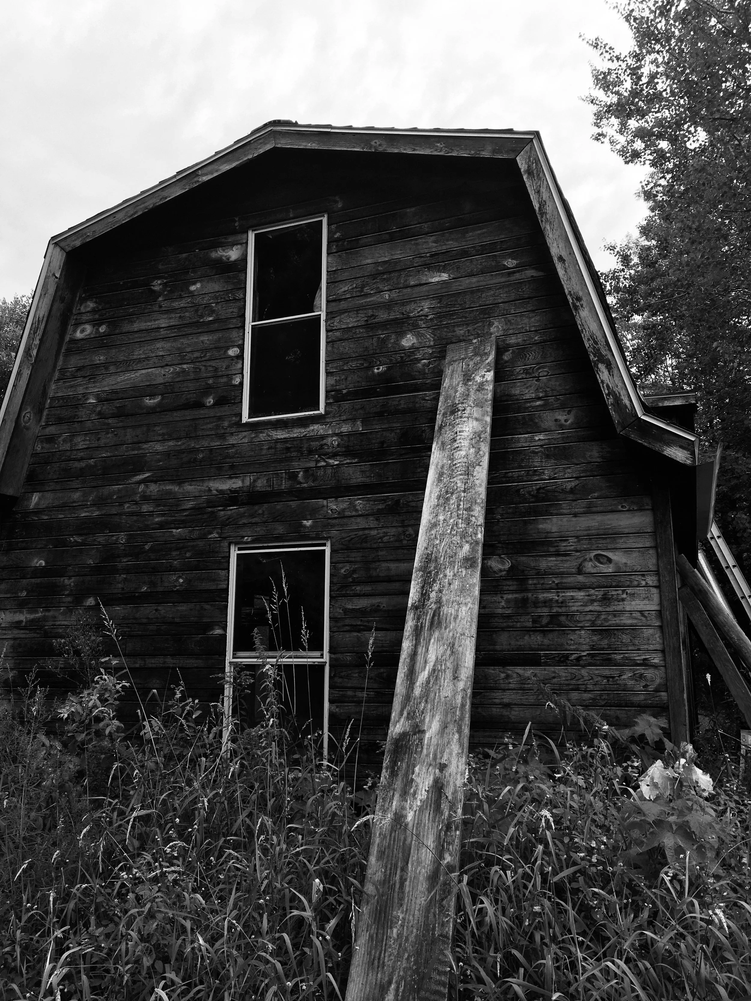 A black and white photo of an old, weathered wooden barn with two vertical windows, surrounded by overgrown grass and trees.