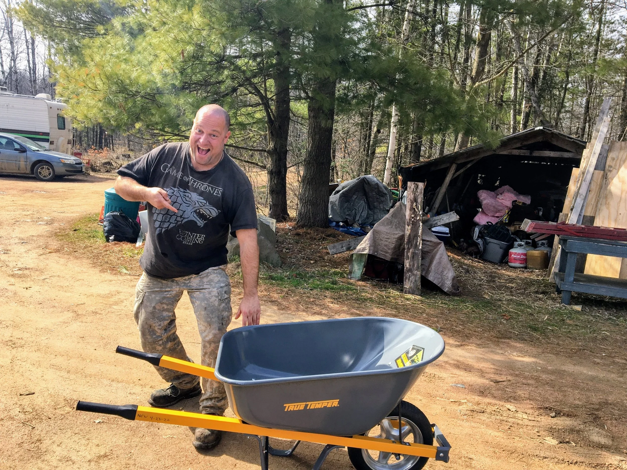 A man with a beard and a Game of Thrones T-shirt smiling and pointing at a wheelbarrow in an outdoor setting with trees, a shed, and parked vehicles in the background.