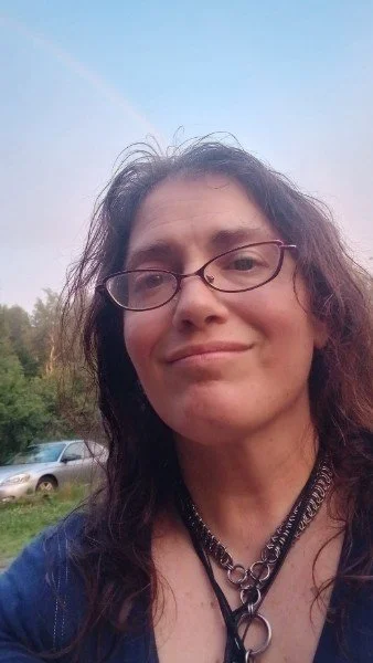 A woman with glasses and dark, curly hair outdoors under a blue sky with a rainbow, standing near parked cars.