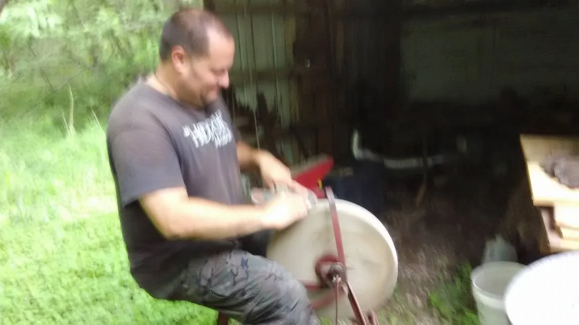 A man sits on a stool outdoors, operating a circular saw or similar tool, with a wooded area and a shed in the background.