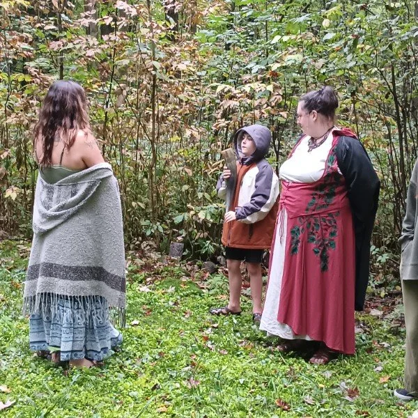 Four people standing outdoors in a wooded area, engaging in a conversation or ritual. Two women and two children, one girl and one boy.