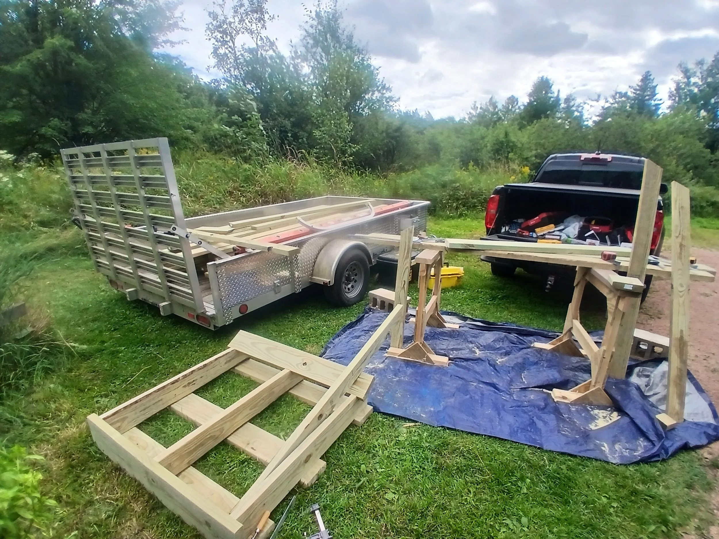 A work-in-progress outdoor woodworking project, with a wooden bench frame on the ground, a partially assembled wooden workbench, and a trailer full of construction materials and tools, set on a grassy area near trees.