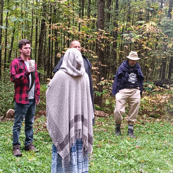 Four people stand outdoors in a forest with green trees and grass, one person is holding a drinking glass, one wears a large hooded shawl, and another wears a hat and beige pants.