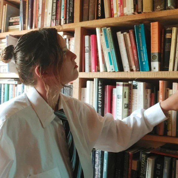 A woman wearing a white shirt and striped tie reaching for a book on a wooden bookshelf in a library.